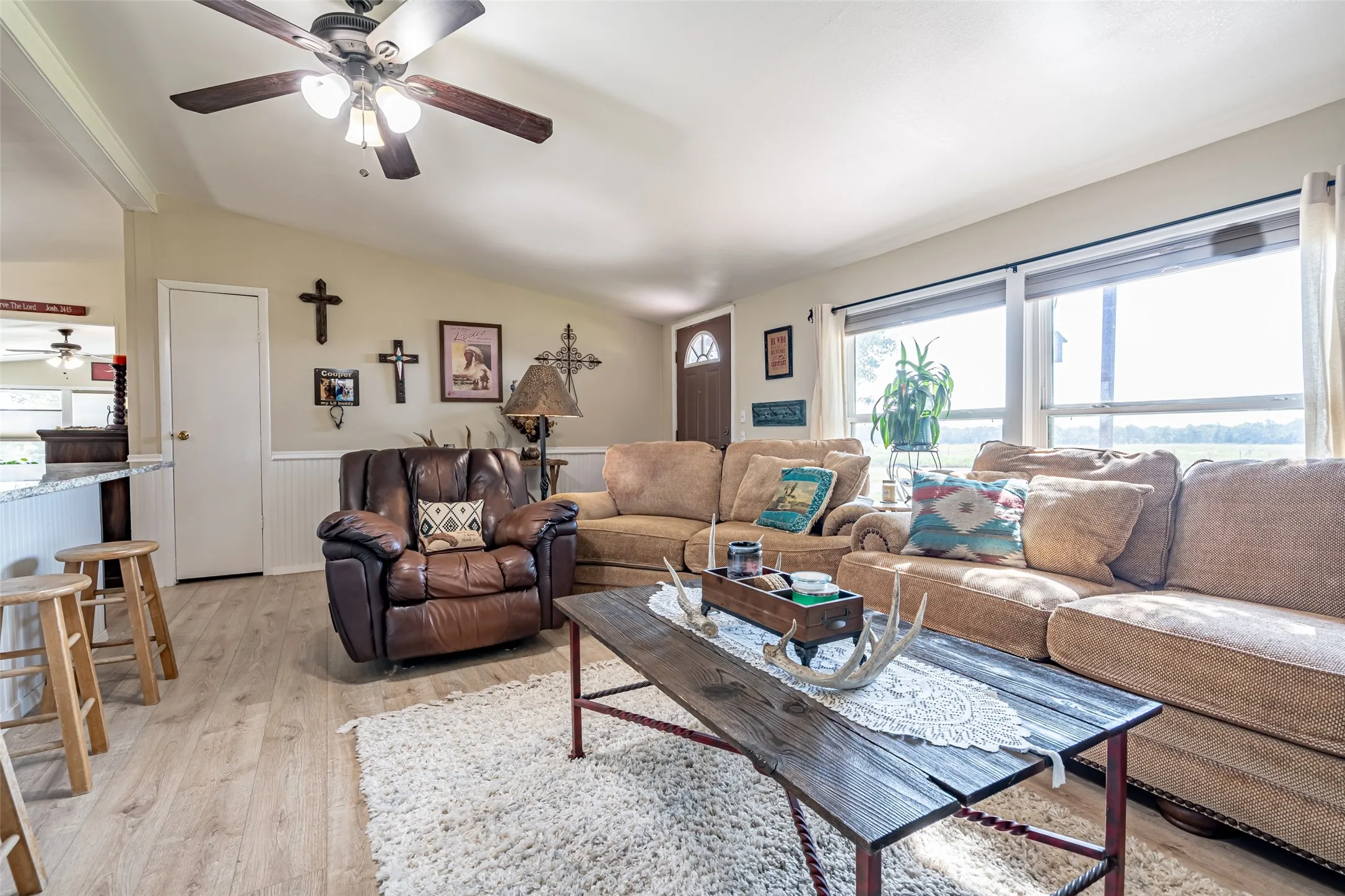Living area with lofted ceiling, healthy amount of natural light, light wood-type flooring, a wainscoted wall, and a ceiling fan.