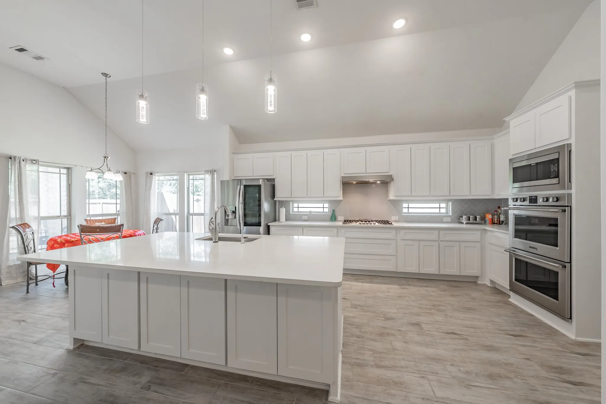 Kitchen with decorative light fixtures, stainless steel appliances, white cabinetry, an island with sink, and high vaulted ceiling