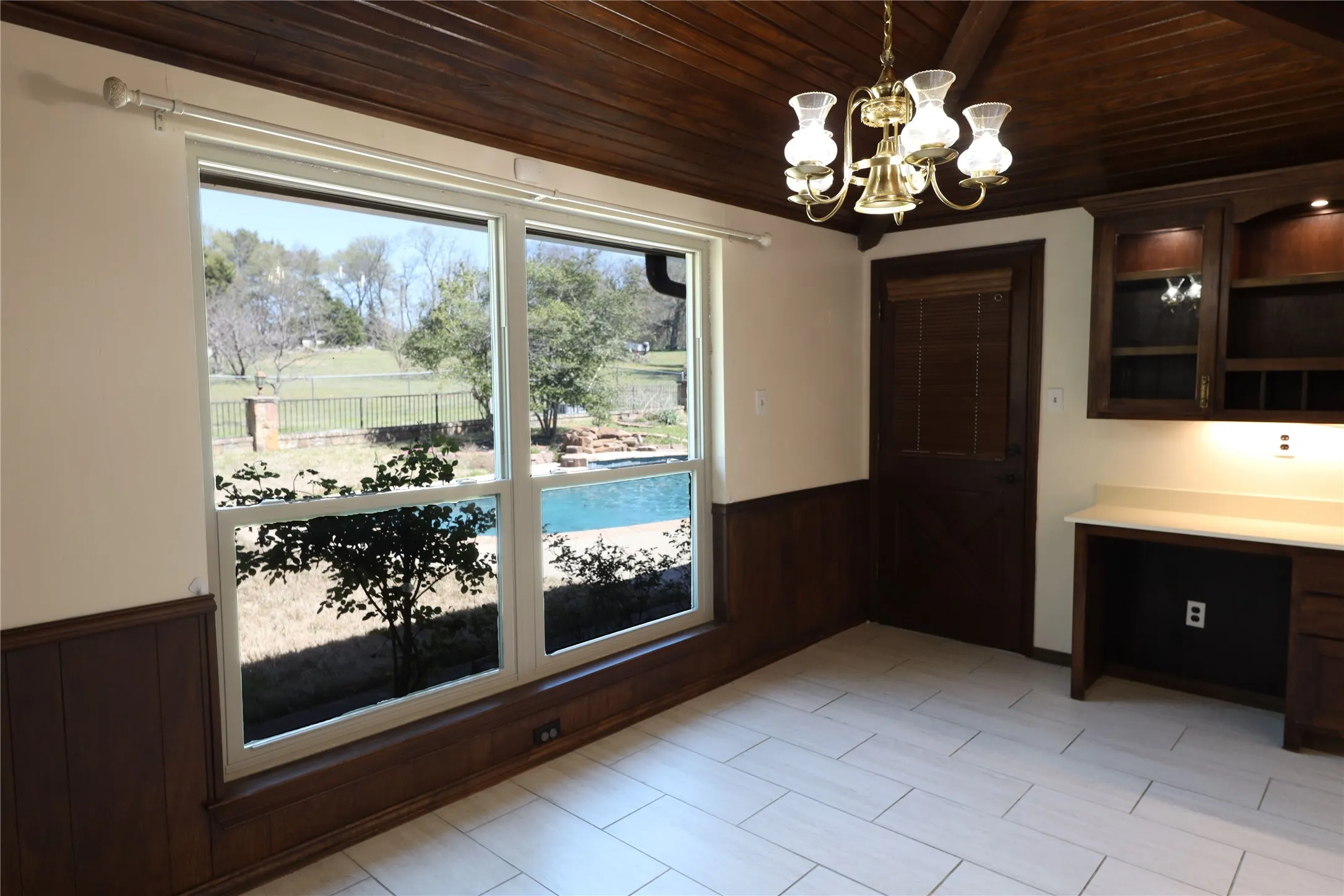 Bright interior space featuring a large window overlooking an outdoor swimming pool, a wood-paneled ceiling, and light-colored tile flooring