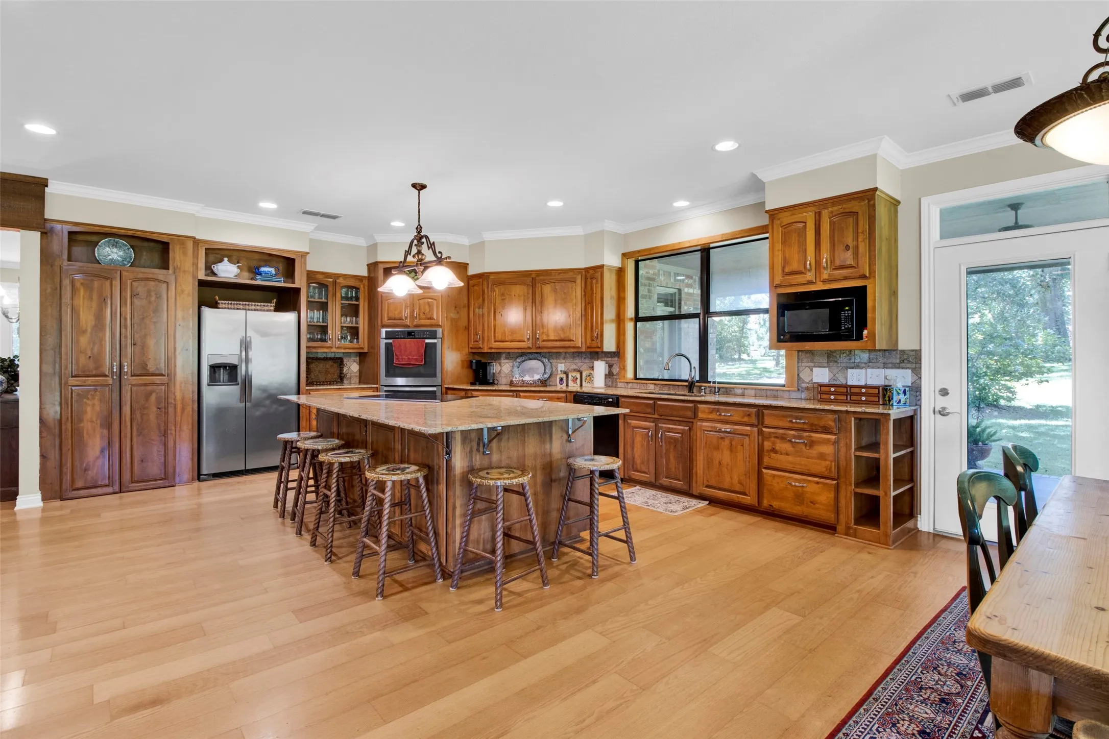 Huge open kitchen featuring a large central island with seating, extensive wood cabinetry, light-toned wood-finish flooring, recessed lighting, and stainless steel appliances, granite countertops. Kitchen has its own dedicated water tank to supply hot water when the house is full!
