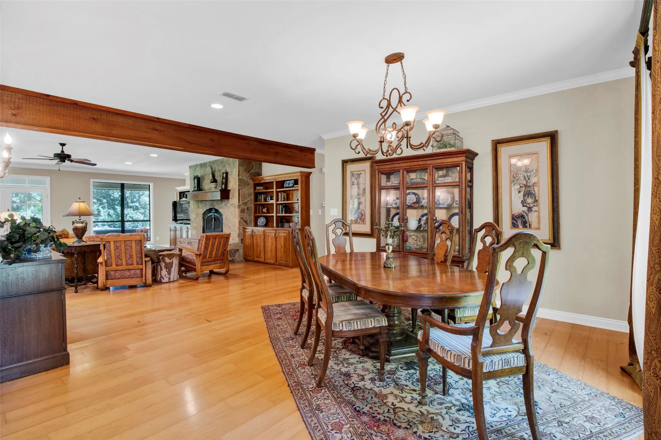 Open-concept dining and living areas featuring wood-finish flooring, a decorative chandelier, crown molding, and a stone-clad fireplace with built-in cabinetry