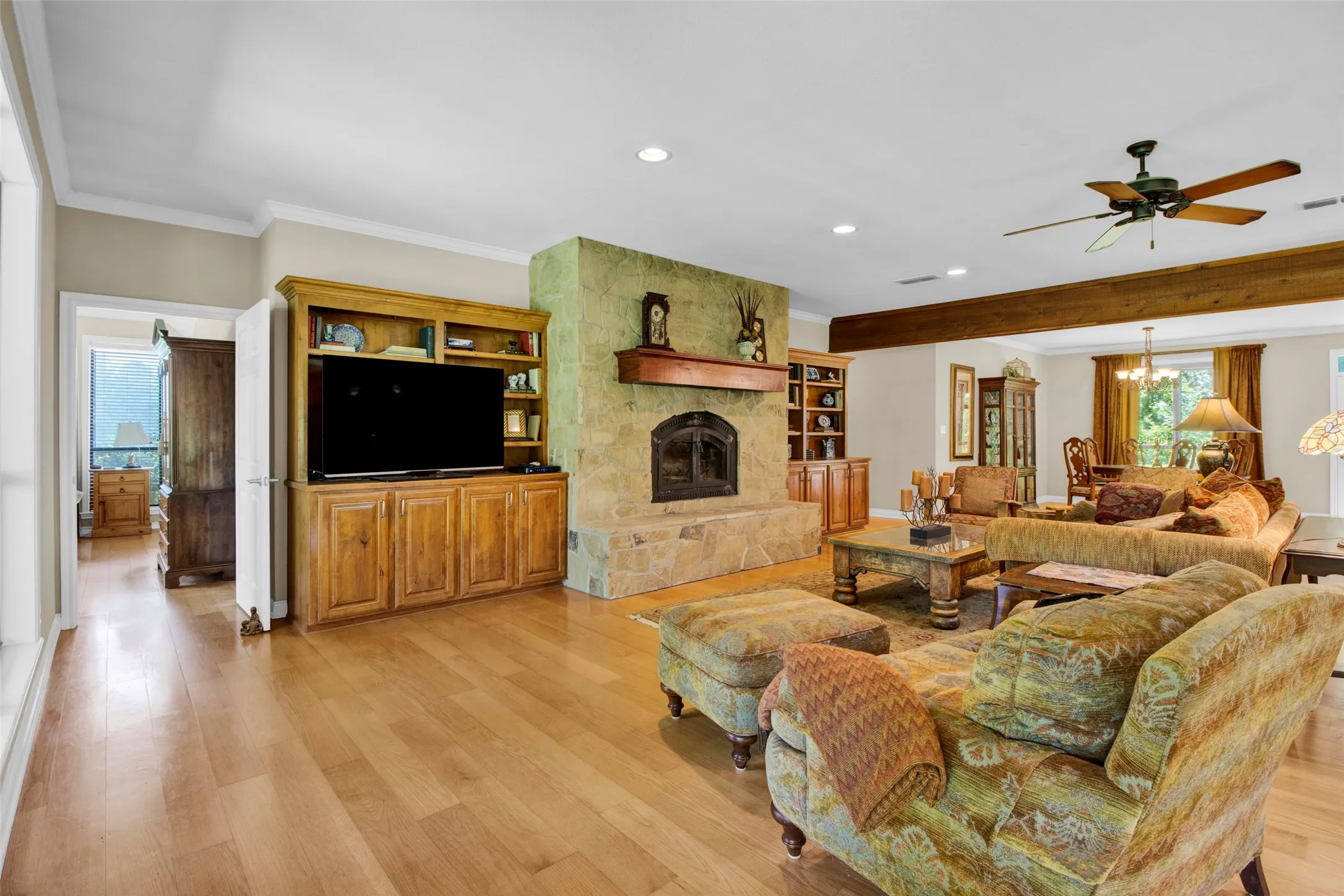 Spacious living area featuring light wood-finish flooring, a stone fireplace with a wood mantle, a built-in entertainment center with storage, a ceiling fan, and a wood beam accent