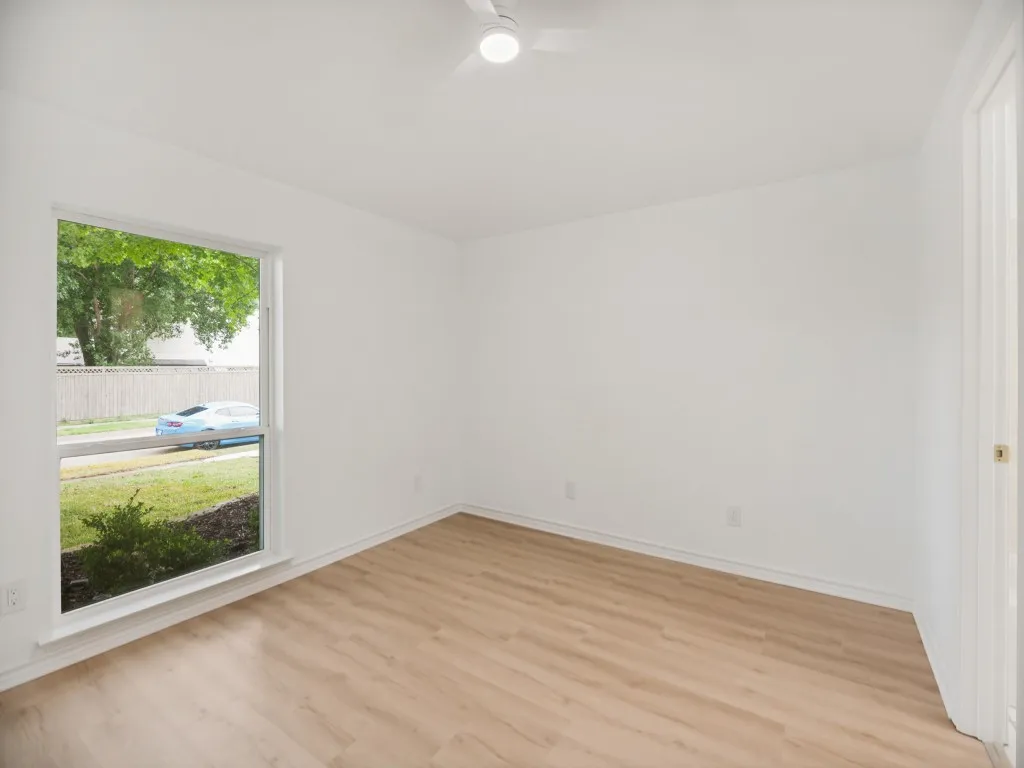 Bright room featuring wood-finish flooring, white walls, a ceiling fan with integrated lighting, and a large window overlooking exterior landscaping