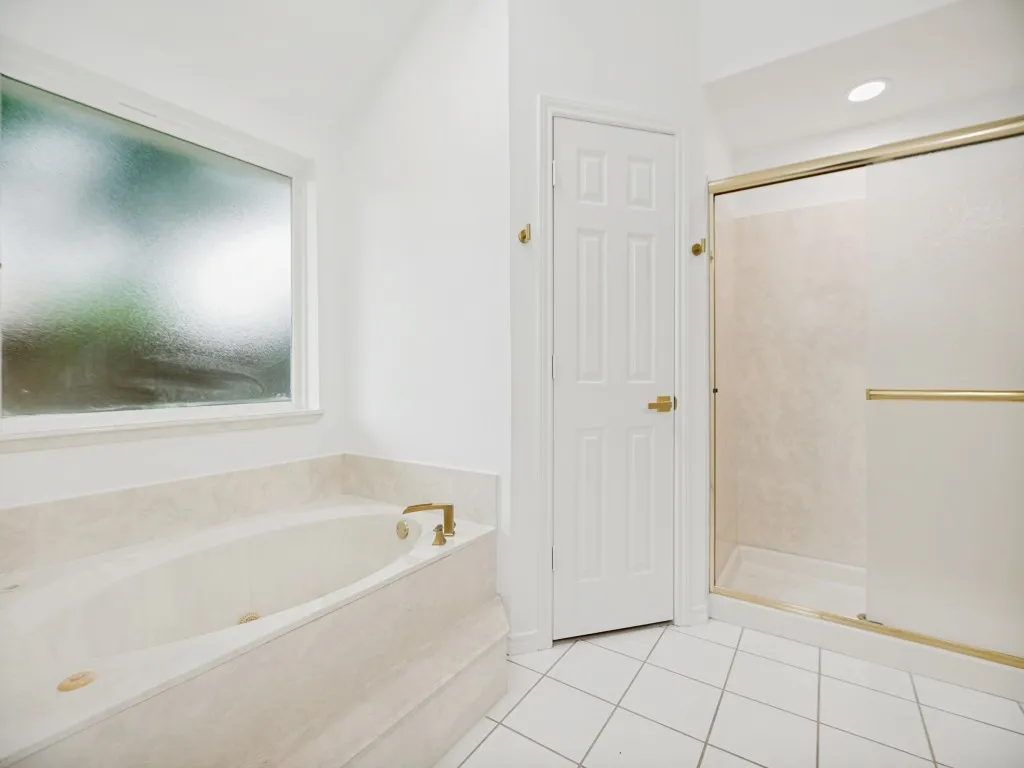 Soaking tub with a tiled surround, frosted window, white paneled door, and a glass-enclosed shower with gold-tone hardware