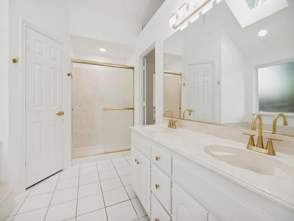 Bathroom featuring a dual vanity with integrated sinks and brass fixtures, a large mirror, a shower with a brass-framed enclosure, white tile flooring, and a skylight