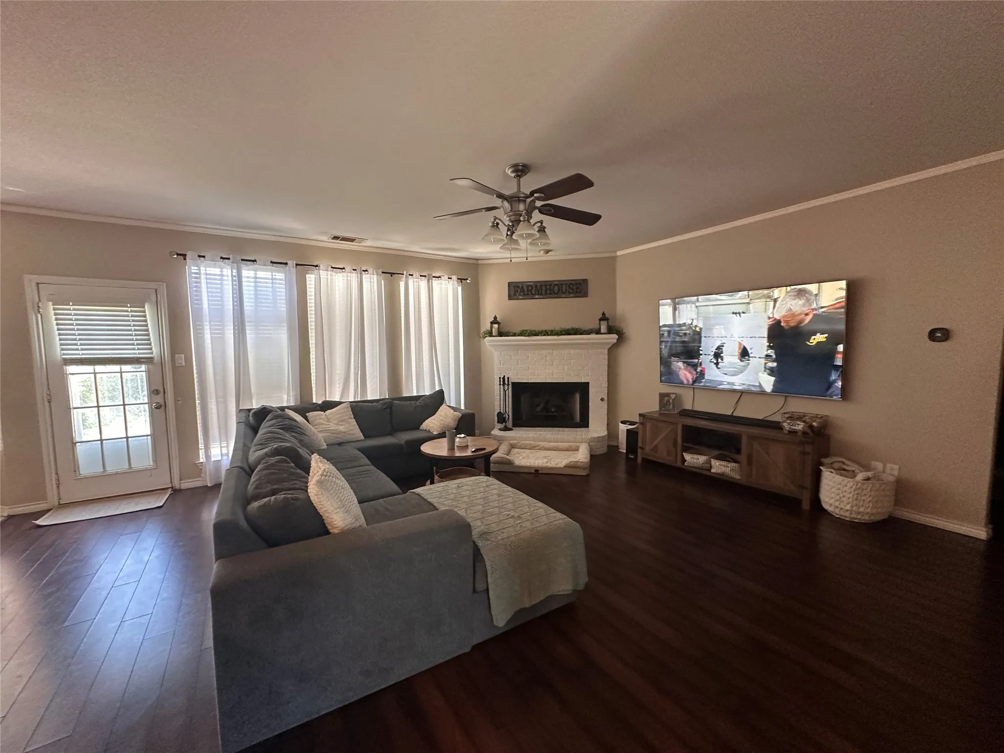 Living area featuring dark wood-finish flooring, a white brick fireplace with a mantel, and a ceiling fan with integrated lighting