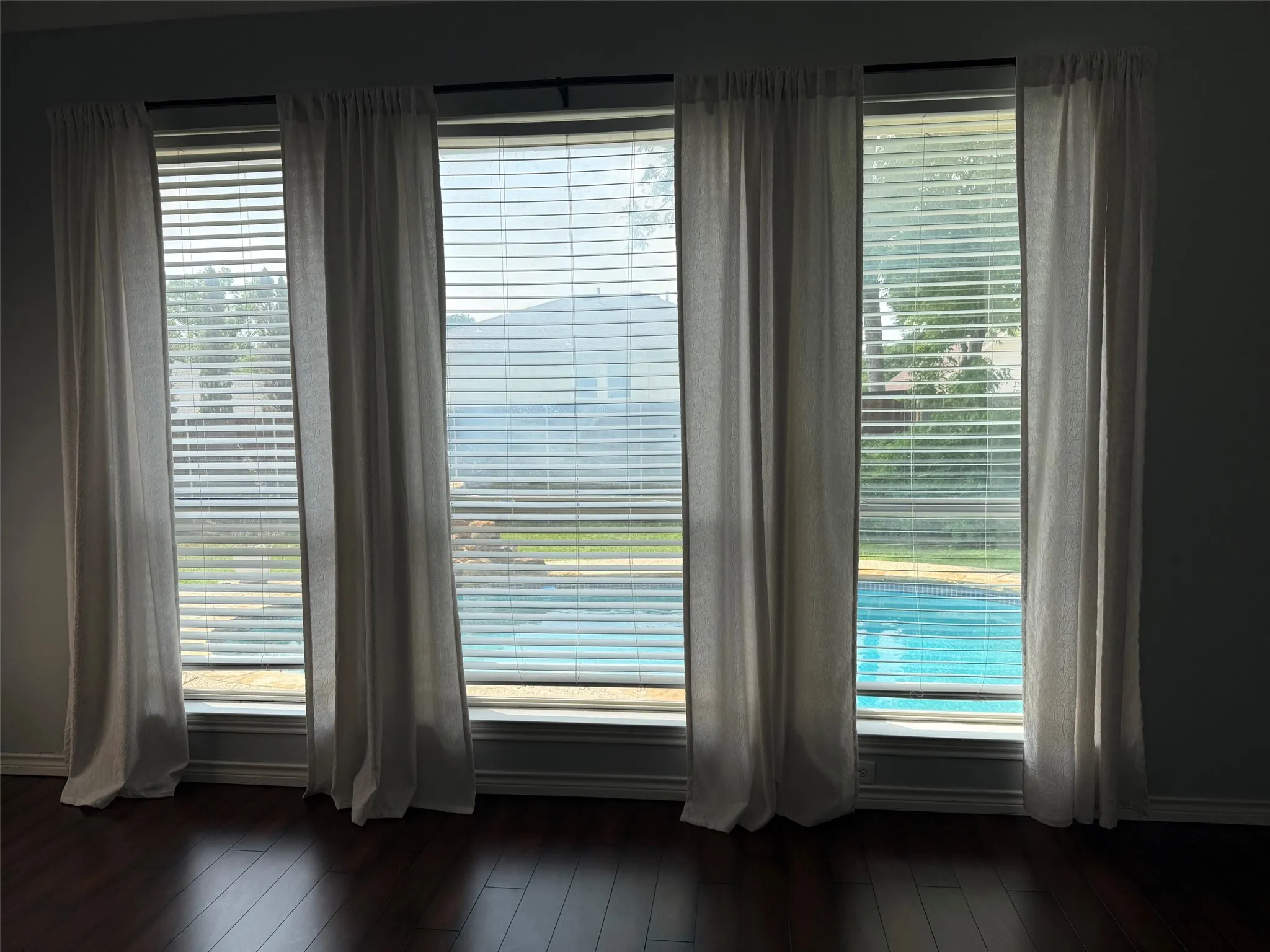 Four large windows overlooking a swimming pool, complete with light-filtering blinds and hanging drapery
