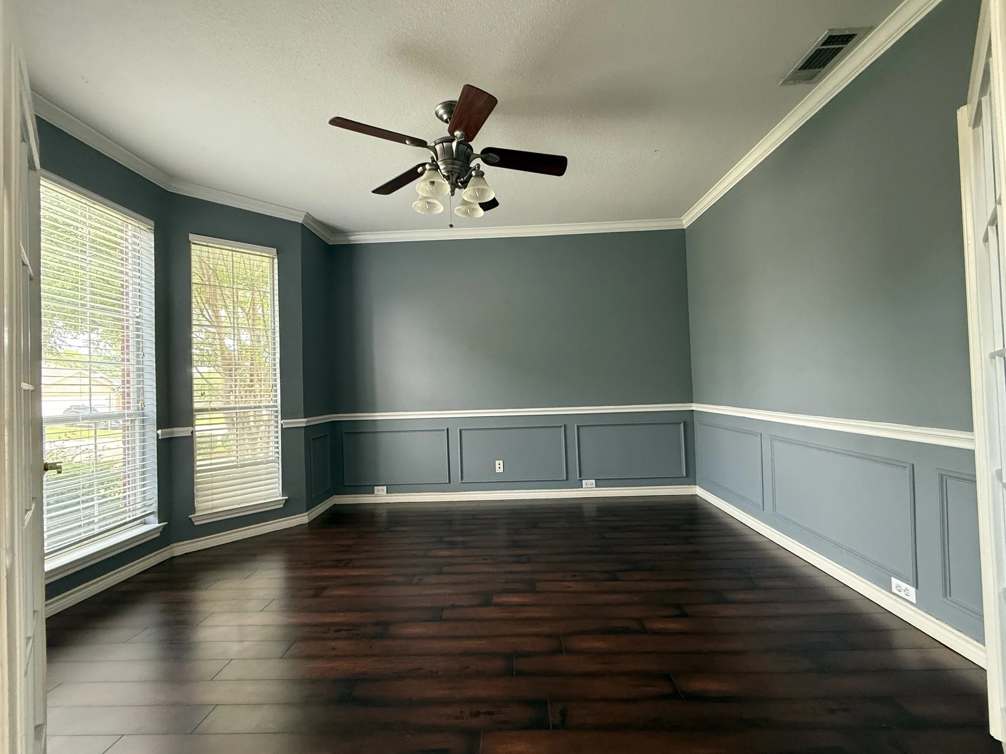 Spacious study room featuring dark wood-finish flooring, wainscoting, crown molding, and a ceiling fan with integrated lighting