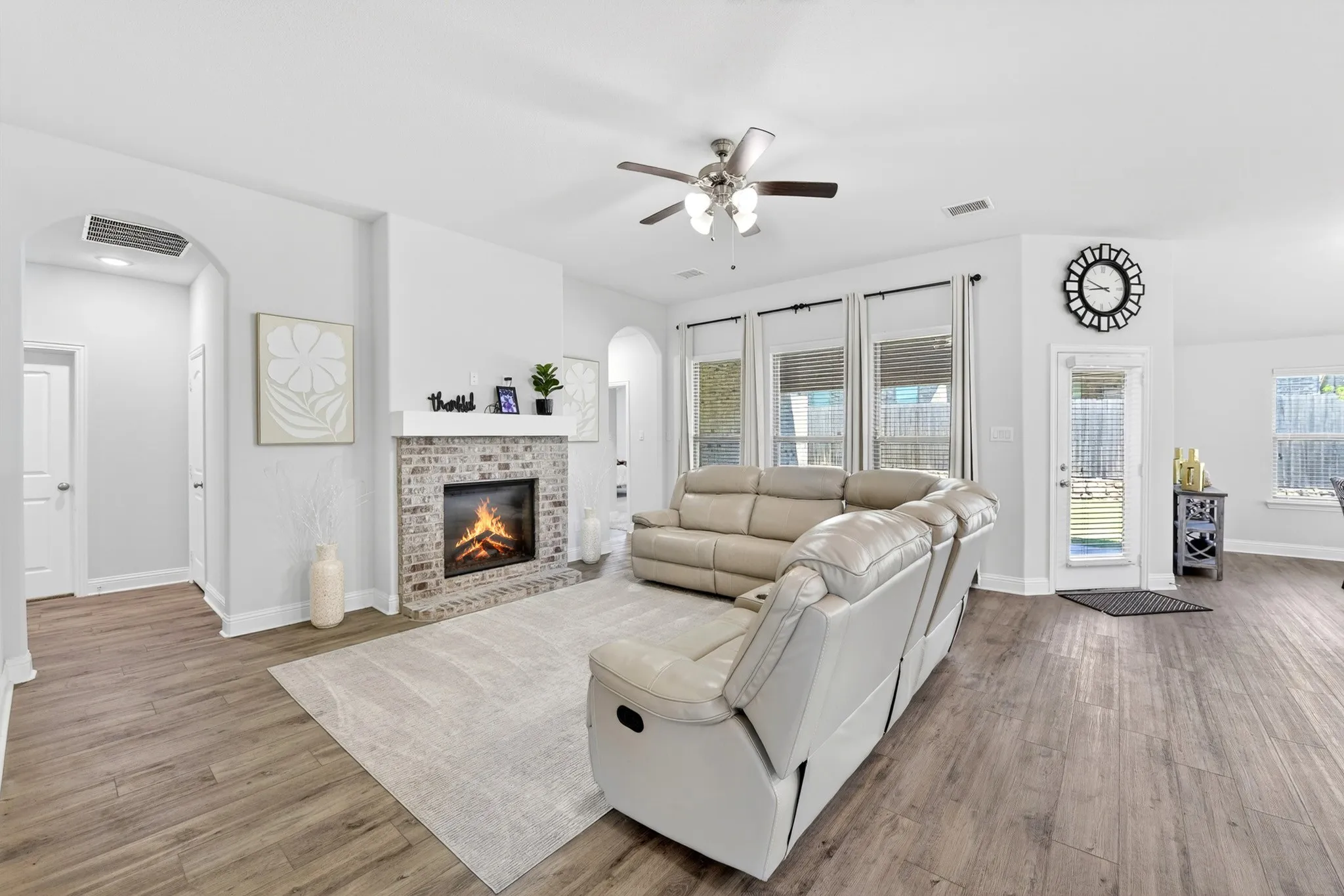Spacious living area featuring wood-finish flooring, a brick-front fireplace with a white mantel, multiple windows, an exterior door, and a ceiling fan