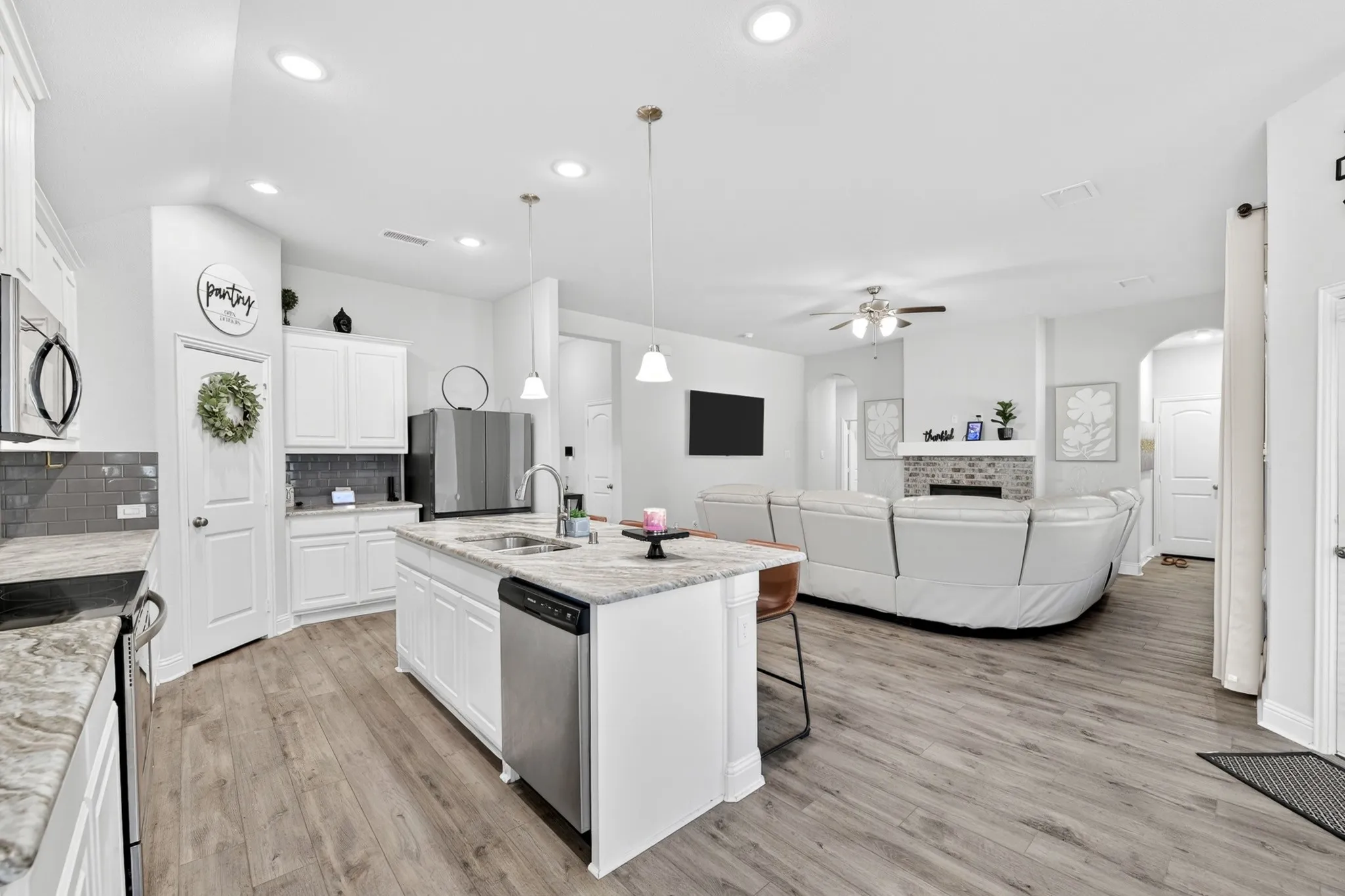 Open-concept living area featuring a kitchen island with integrated sink, wood-finish flooring, recessed lighting, white cabinetry, and a grey subway tile backsplash