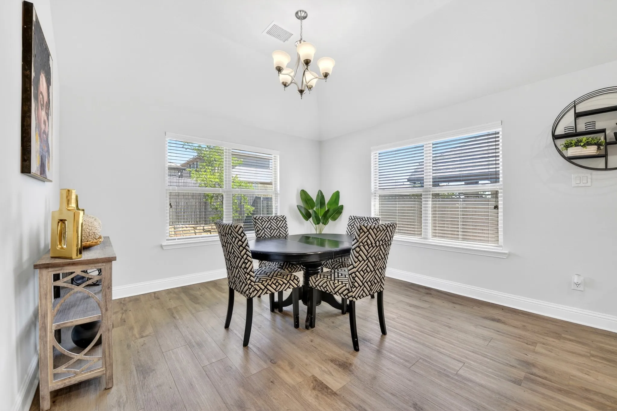 Spacious dining area featuring wood-finish flooring, two large windows with blinds, a vaulted ceiling, and an overhead chandelier with frosted glass shades