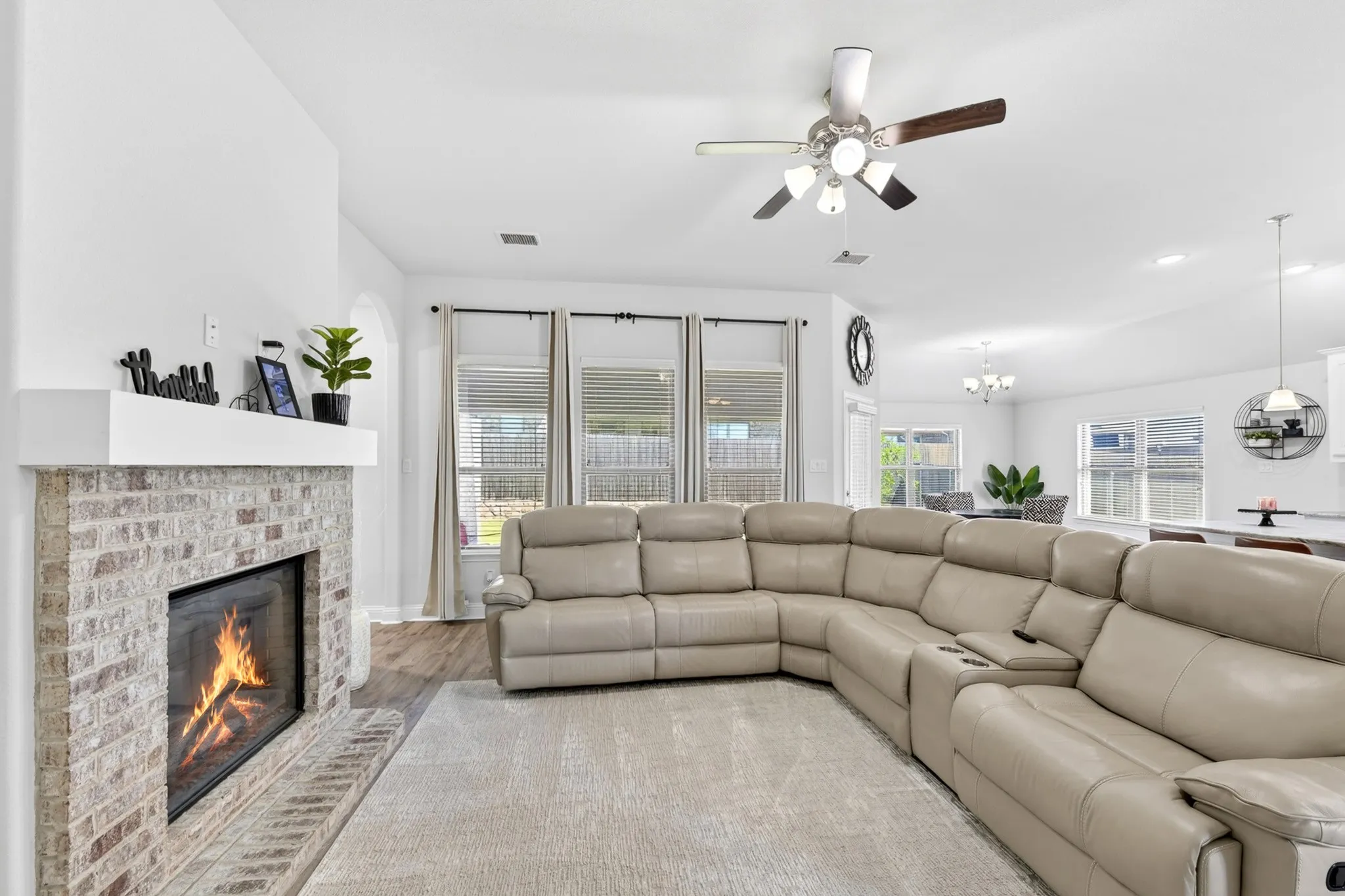 Living area featuring a brick fireplace with a white mantle, wood-finish flooring, a ceiling fan with light fixture, and multiple windows with blinds