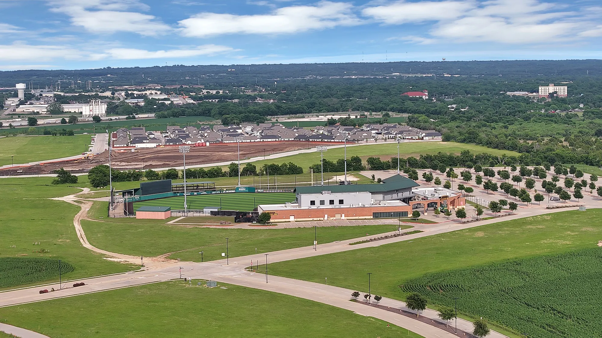 Aerial perspective showcasing extensive green spaces, a baseball field with stadium seating and field lighting, a large adjacent parking facility, and surrounding development