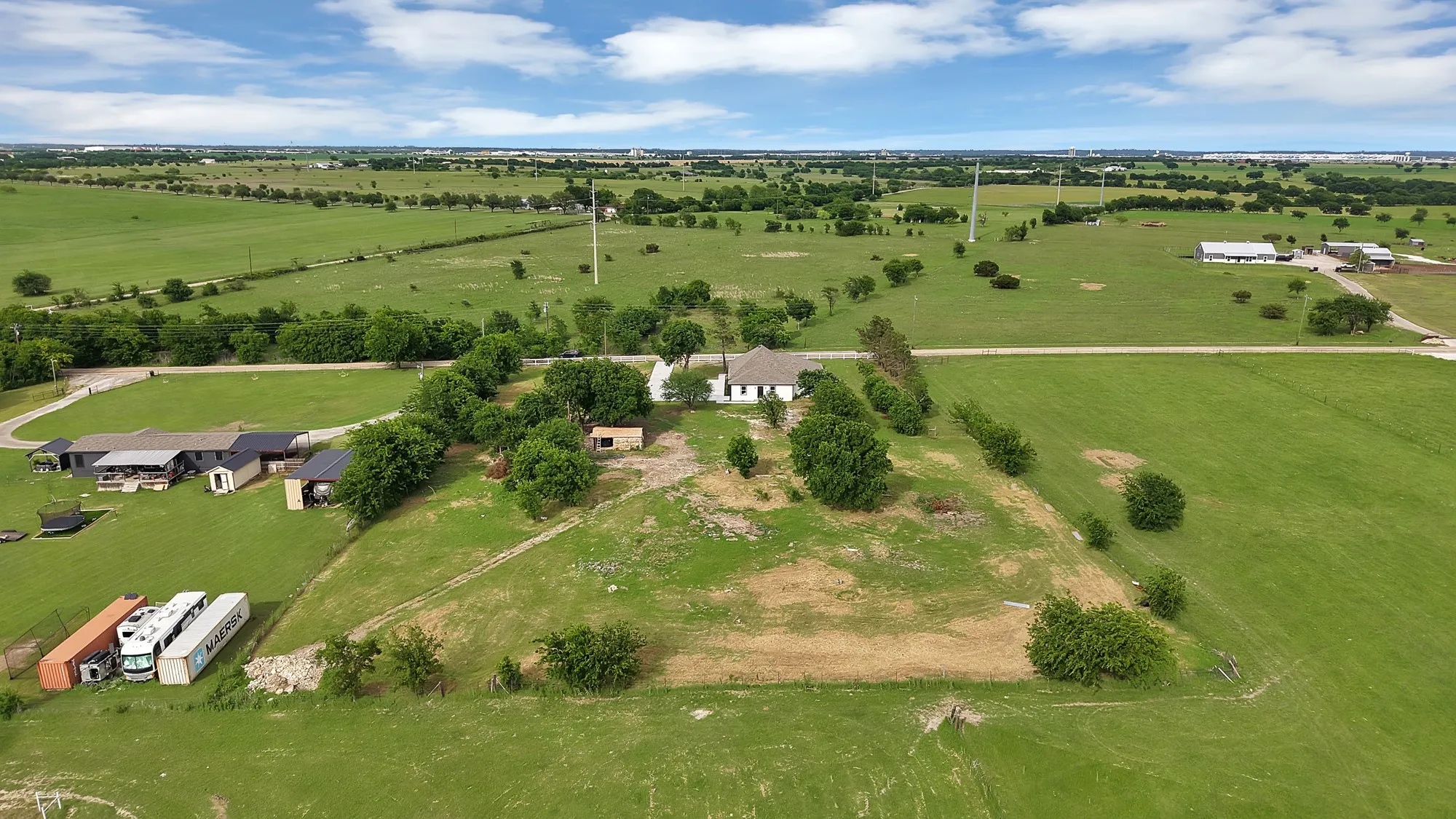 Expansive rural property featuring a single-story residence with a light-colored roof, multiple outbuildings, mature trees, and vast green fields