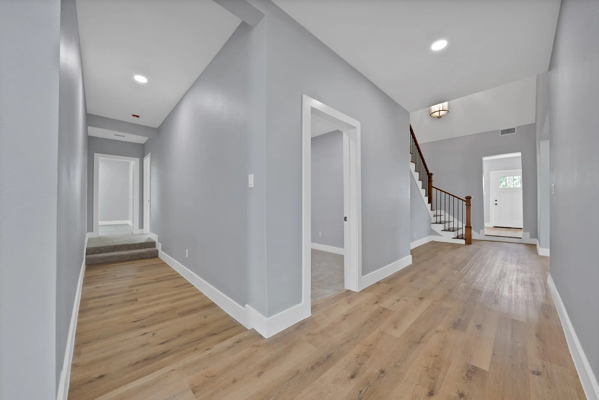 Spacious interior hallway featuring wood-finish flooring, light gray walls, white trim, recessed lighting, and a staircase with dark wood handrails and black metal balusters