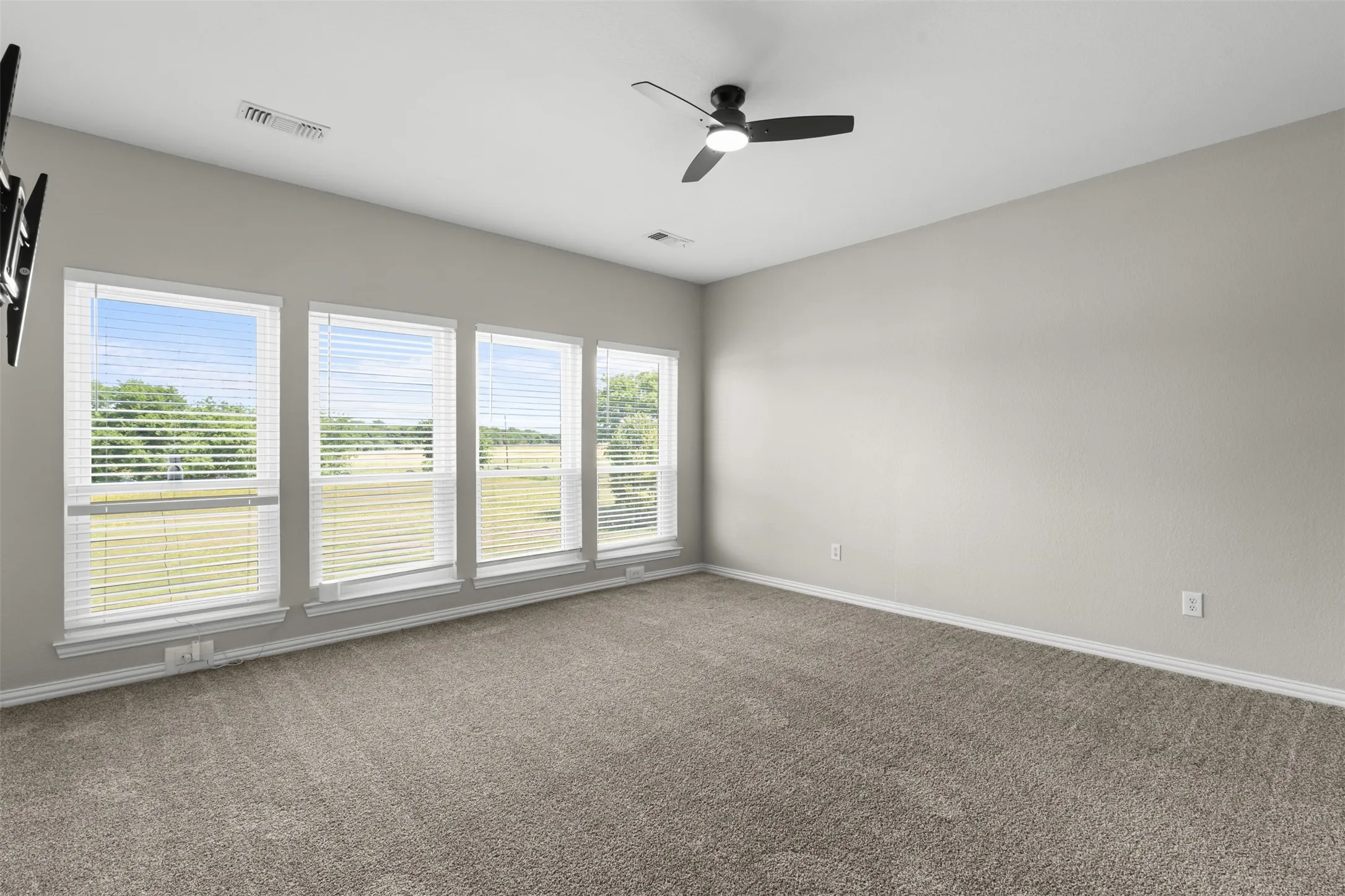Spacious room featuring four elongated windows with blinds, a wall-mounted ceiling fan, and grey carpet flooring