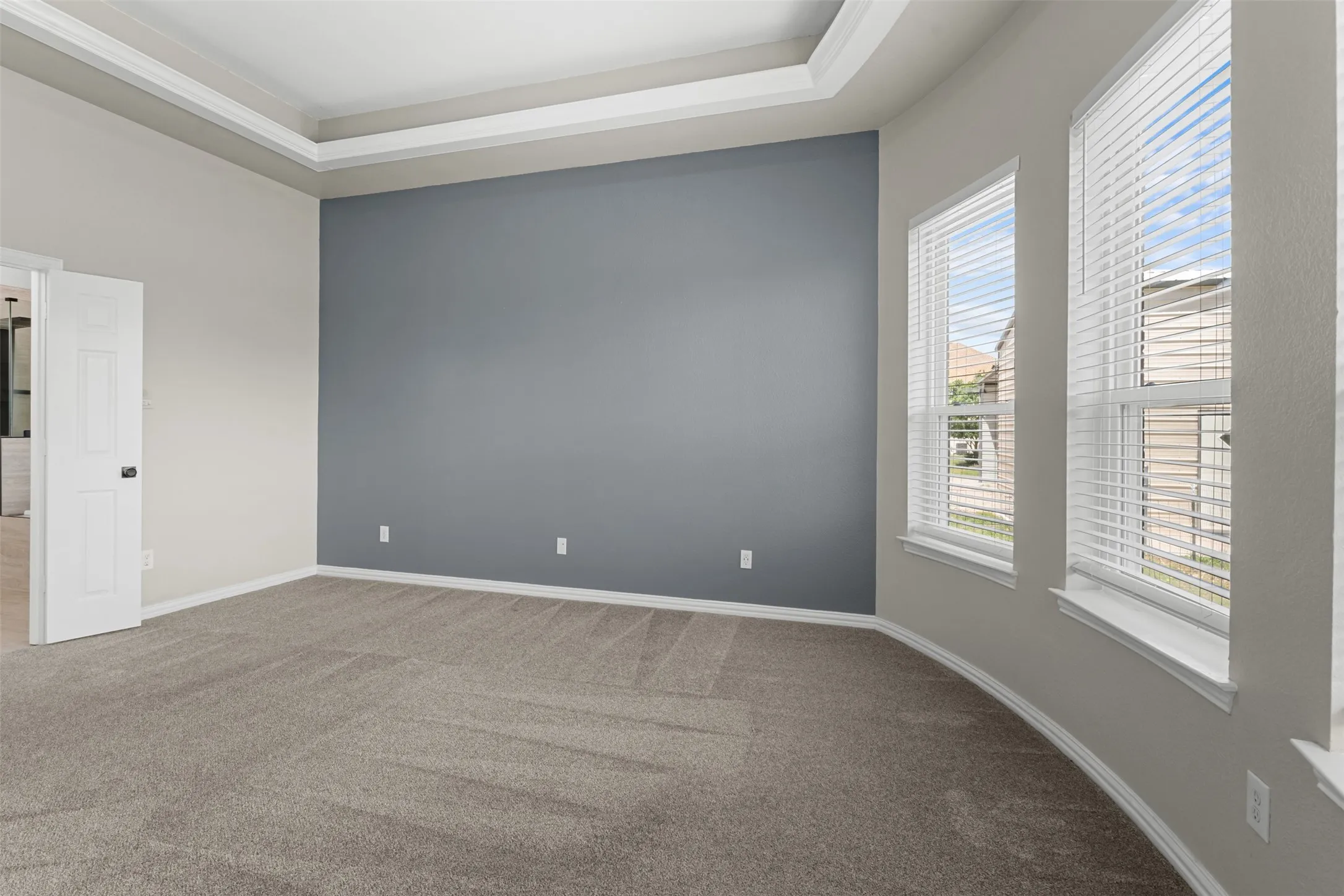 Spacious room featuring tray ceilings, a contrasting accent wall, neutral carpeting, and a bay window with blinds