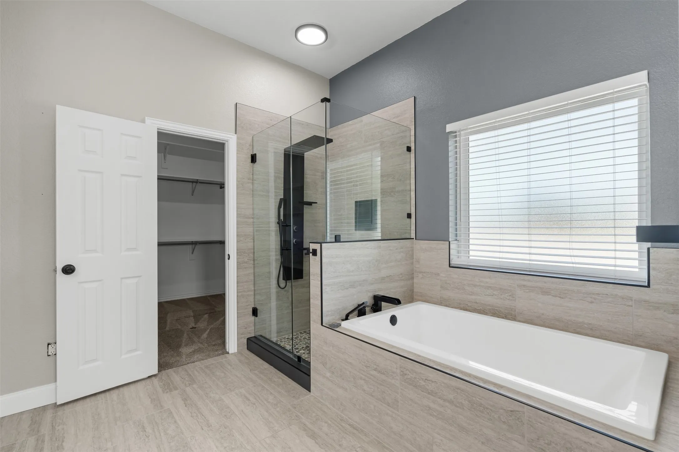 Contemporary master bathroom featuring a freestanding soaking tub, a frameless glass-enclosed shower with black hardware, and light-toned tile flooring