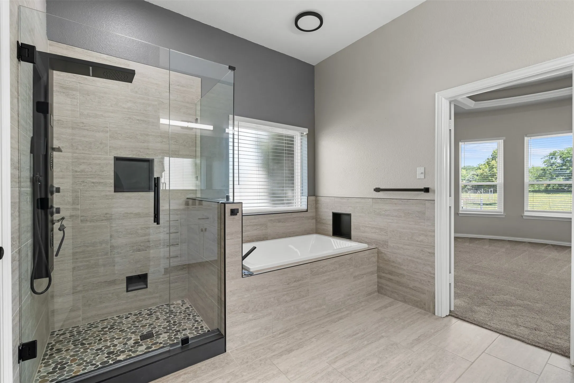 Spacious master bathroom featuring a glass-enclosed shower with pebble flooring and dark hardware, a built-in soaking tub, and large format tile flooring