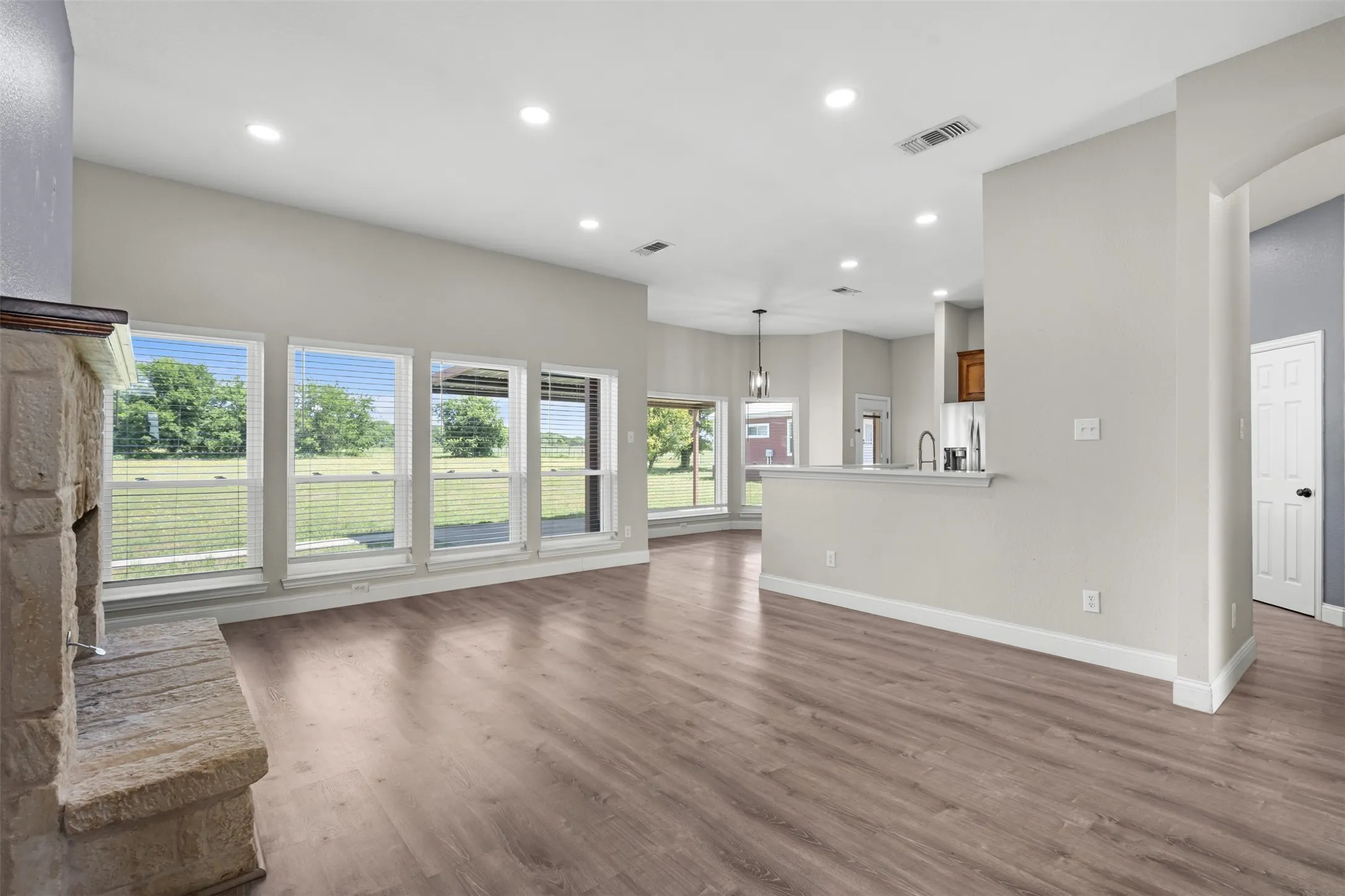 Spacious living area featuring wood-finish flooring, a stone-clad fireplace, recessed lighting, and multiple windows with horizontal blinds