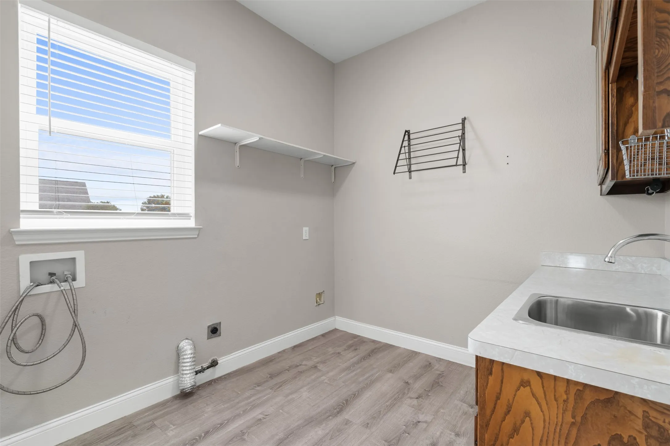 Laundry room featuring a window with blinds, a utility sink with a faucet, wood-finish flooring, and white baseboards