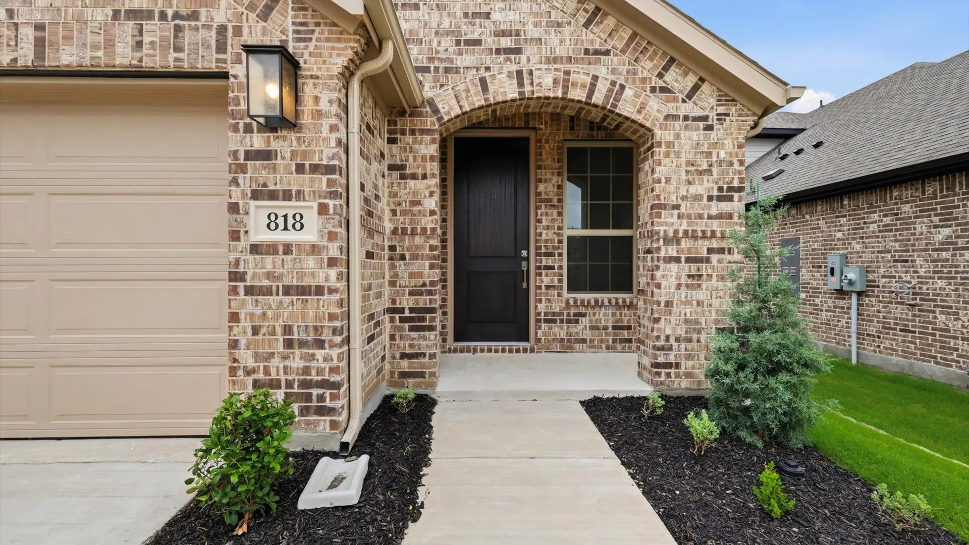 Brick exterior featuring an arched entryway, a dark paneled front door, an exterior wall lantern, a concrete pathway, and a tan garage door
