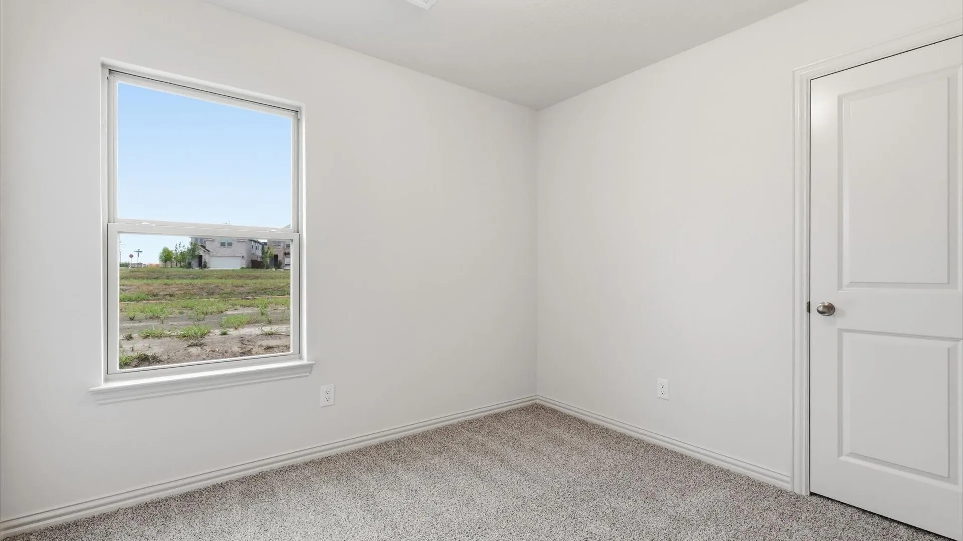 Bright interior room featuring a large single-hung window, light gray carpet flooring, white painted walls, white baseboards, and a white paneled door with a brushed nickel knob