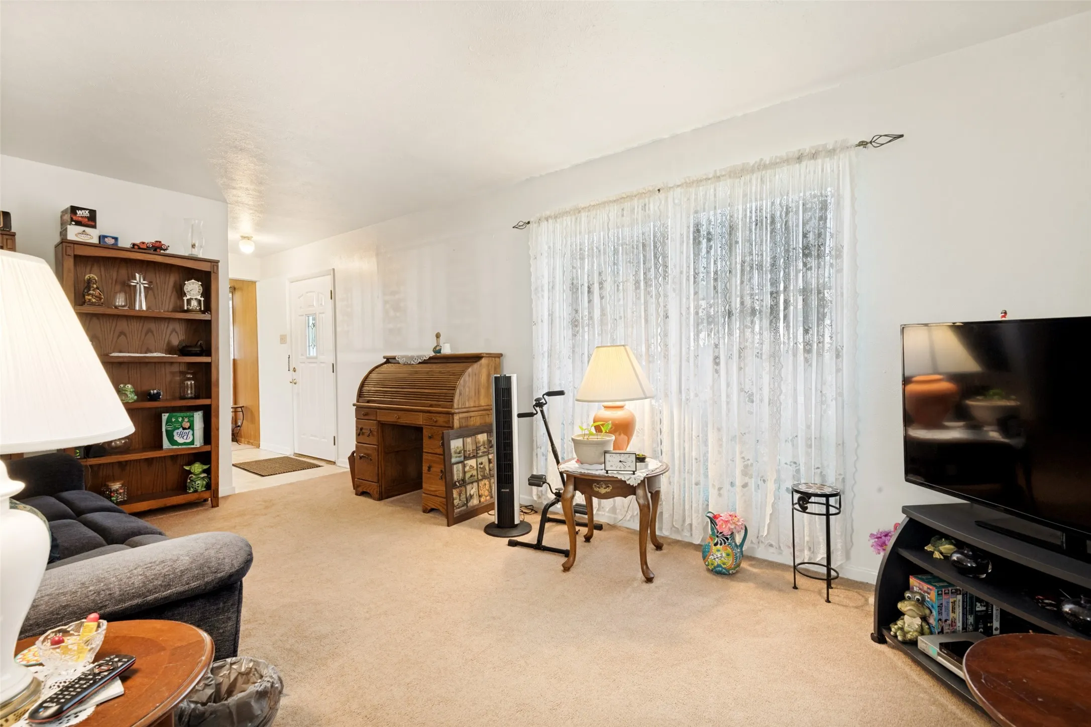 Carpeted living area featuring a large window with sheer curtains, a built-in bookcase, and a white entry door