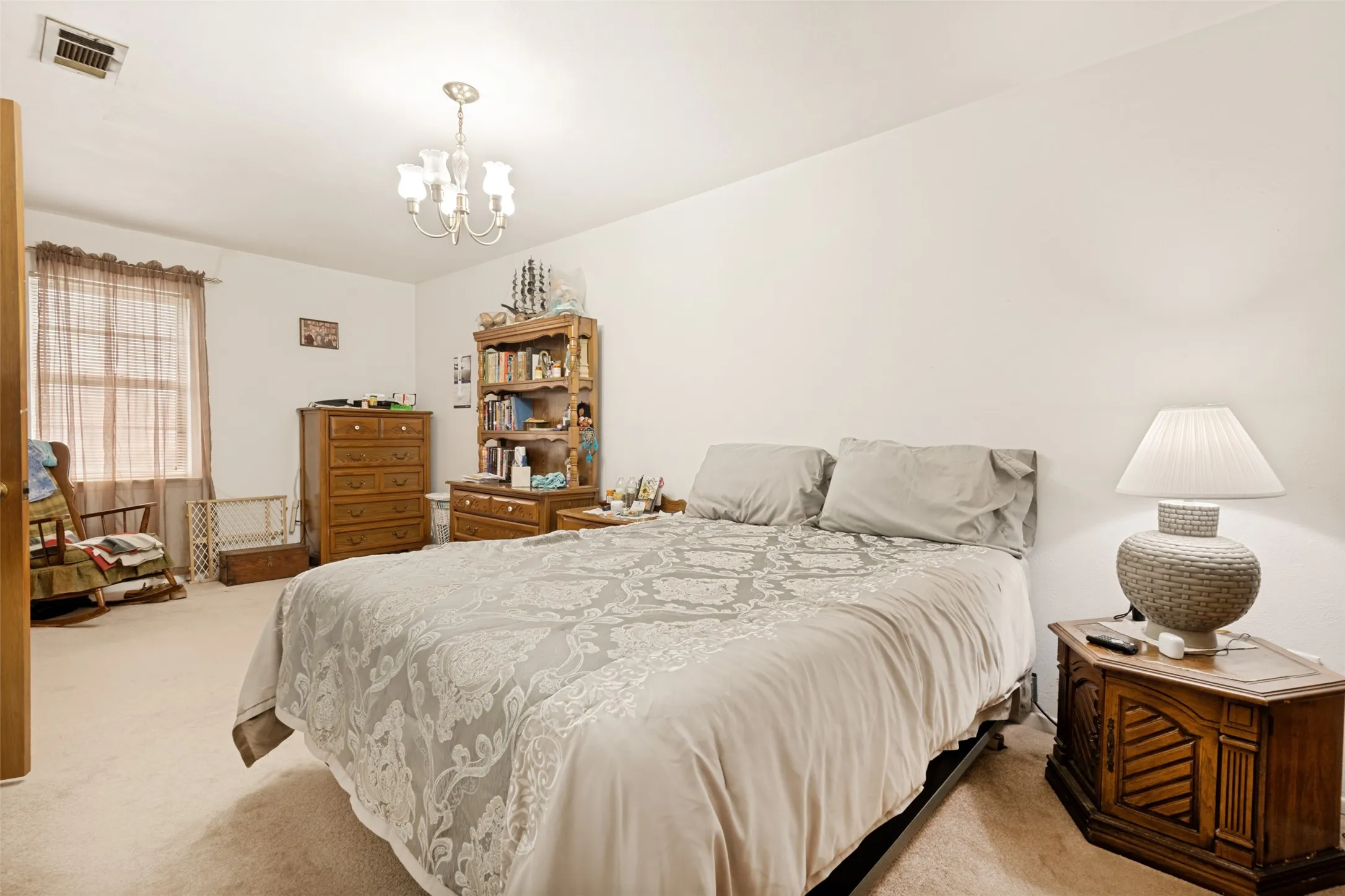 Carpeted bedroom featuring a ceiling-mounted chandelier, window with privacy blinds, and a built-in vent