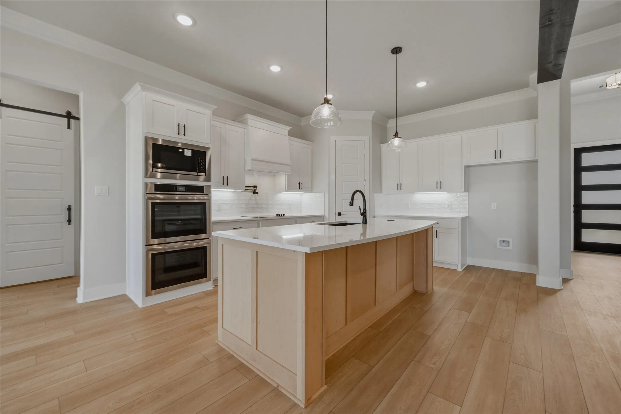 Modern kitchen featuring white shaker-style cabinetry, a warm wood-finish island with seating, stainless steel double wall ovens, quartz countertops, pendant lighting, and wide-plank wood-finish flooring throughout the open concept space