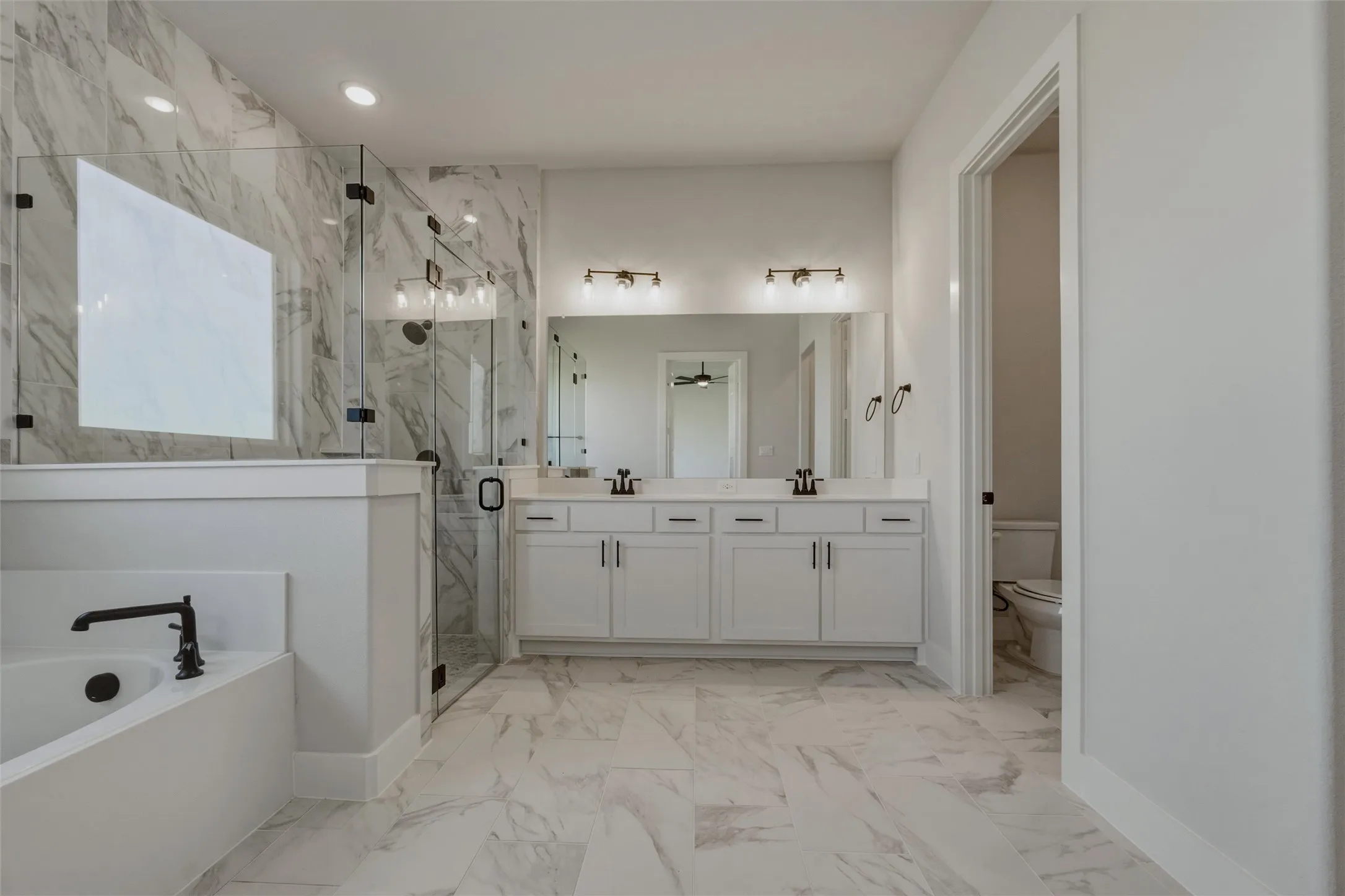 Spacious bathroom featuring a dual vanity with white cabinetry, a glass-enclosed shower, a built-in soaking tub with a matte black fixture, and large-format tile flooring