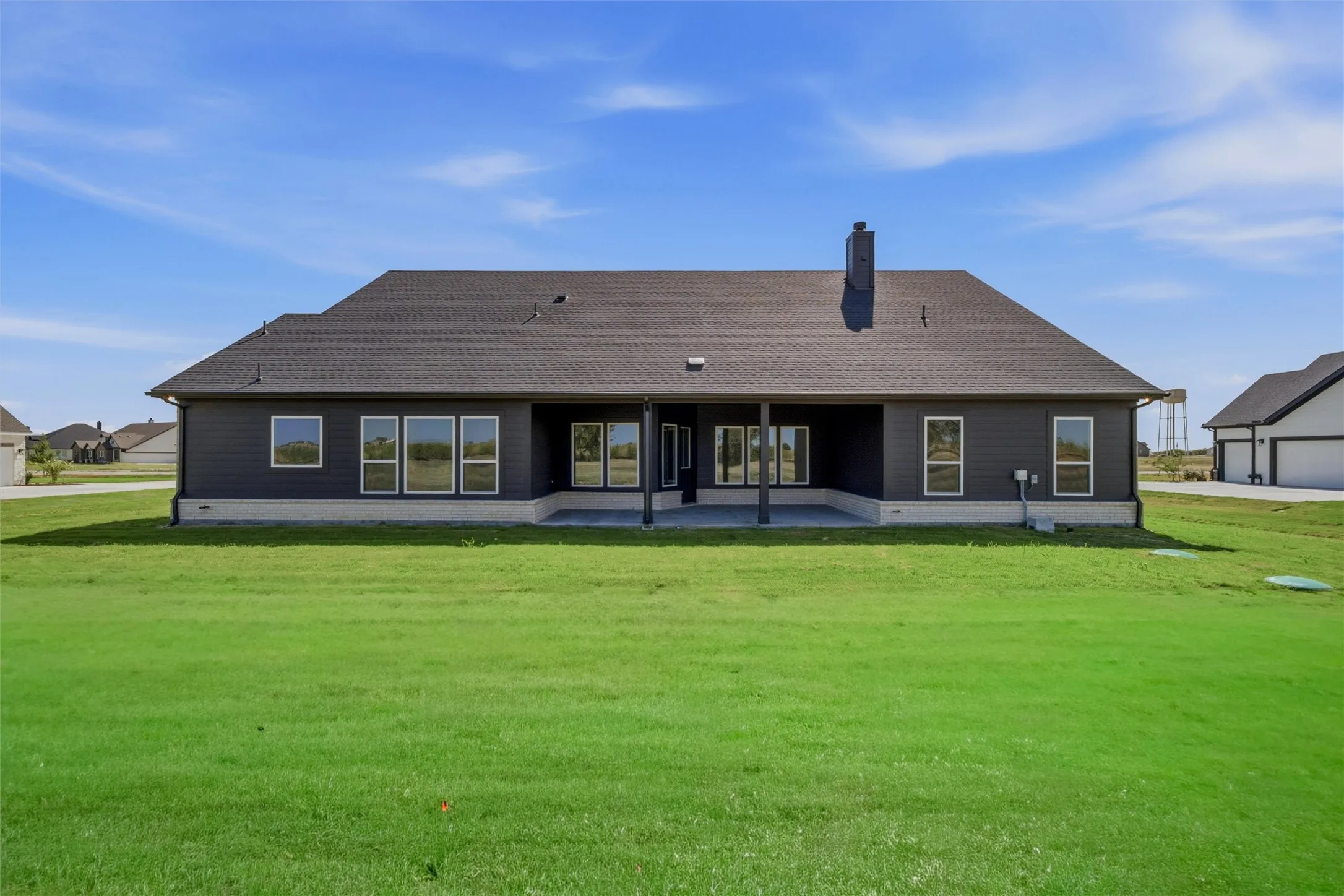 Expansive green yard with a dark shingled roof, dark siding, multiple windows, and a covered concrete patio