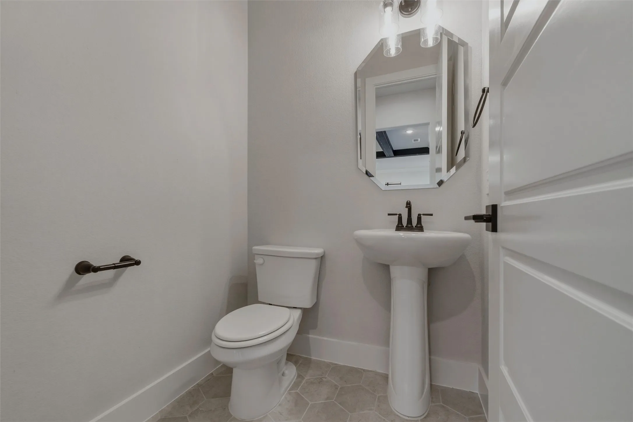 Pedestal sink with dark bronze fixtures, octagonal mirror, and light-toned hexagonal tile flooring