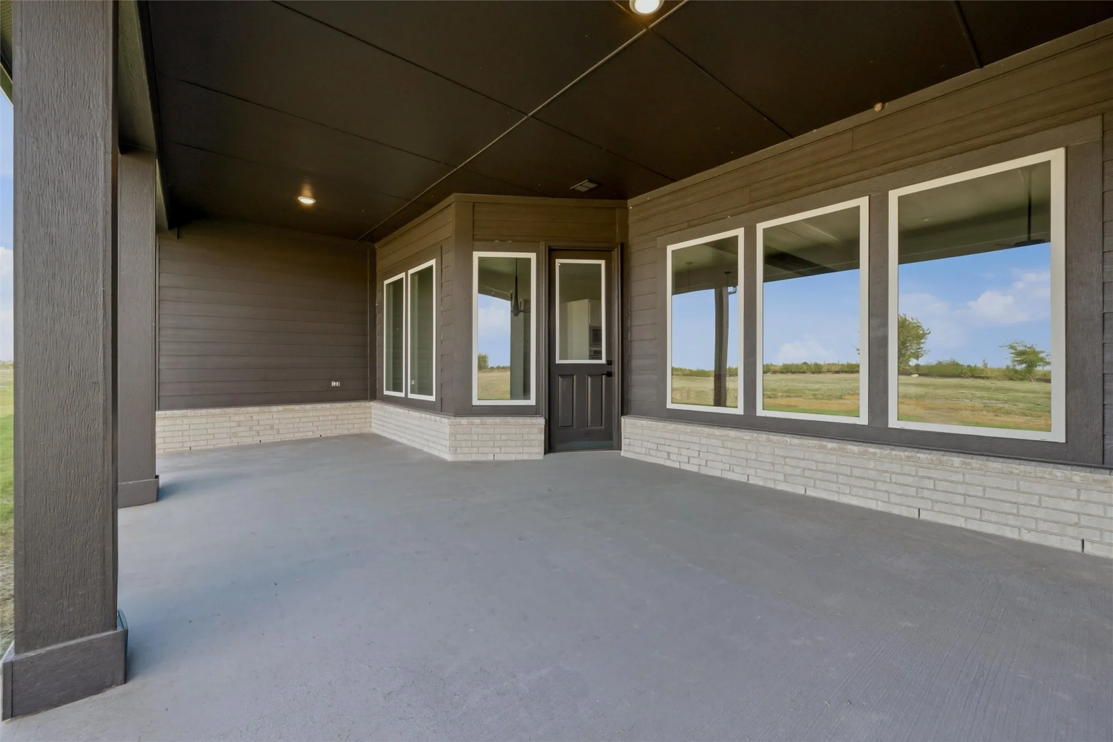 Spacious covered patio with a textured concrete surface, framed by a dark plank siding exterior and a light brick wainscoting