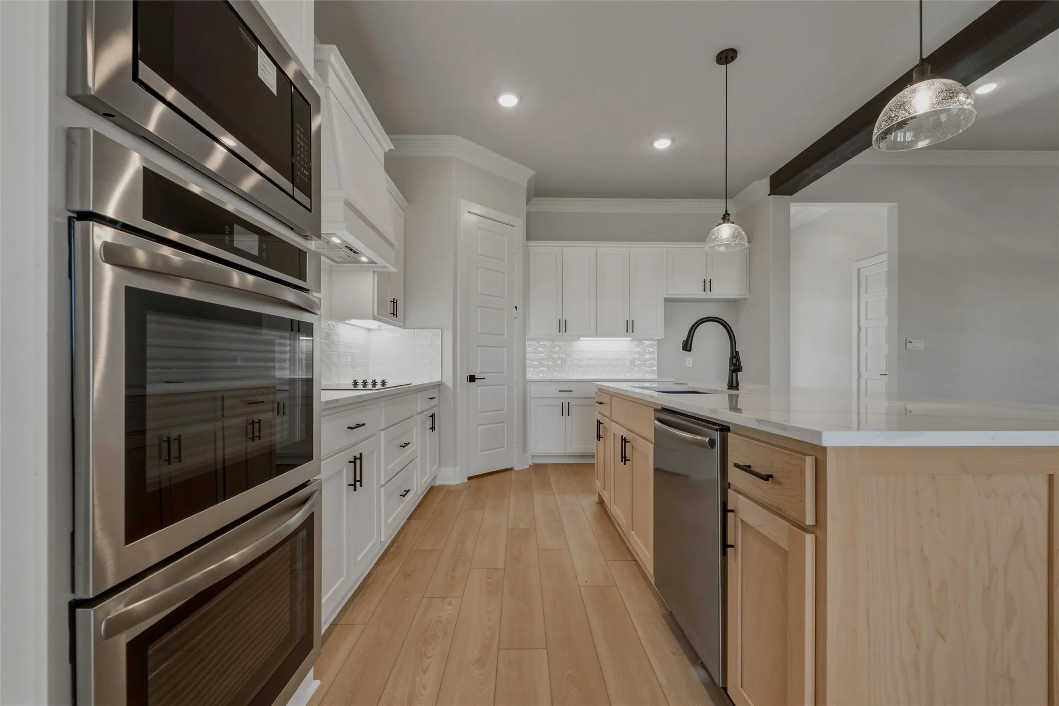 Stainless steel wall oven and microwave, white shaker cabinetry with black hardware, wood-finish flooring, light-toned kitchen island with a built-in sink, and pendant lighting