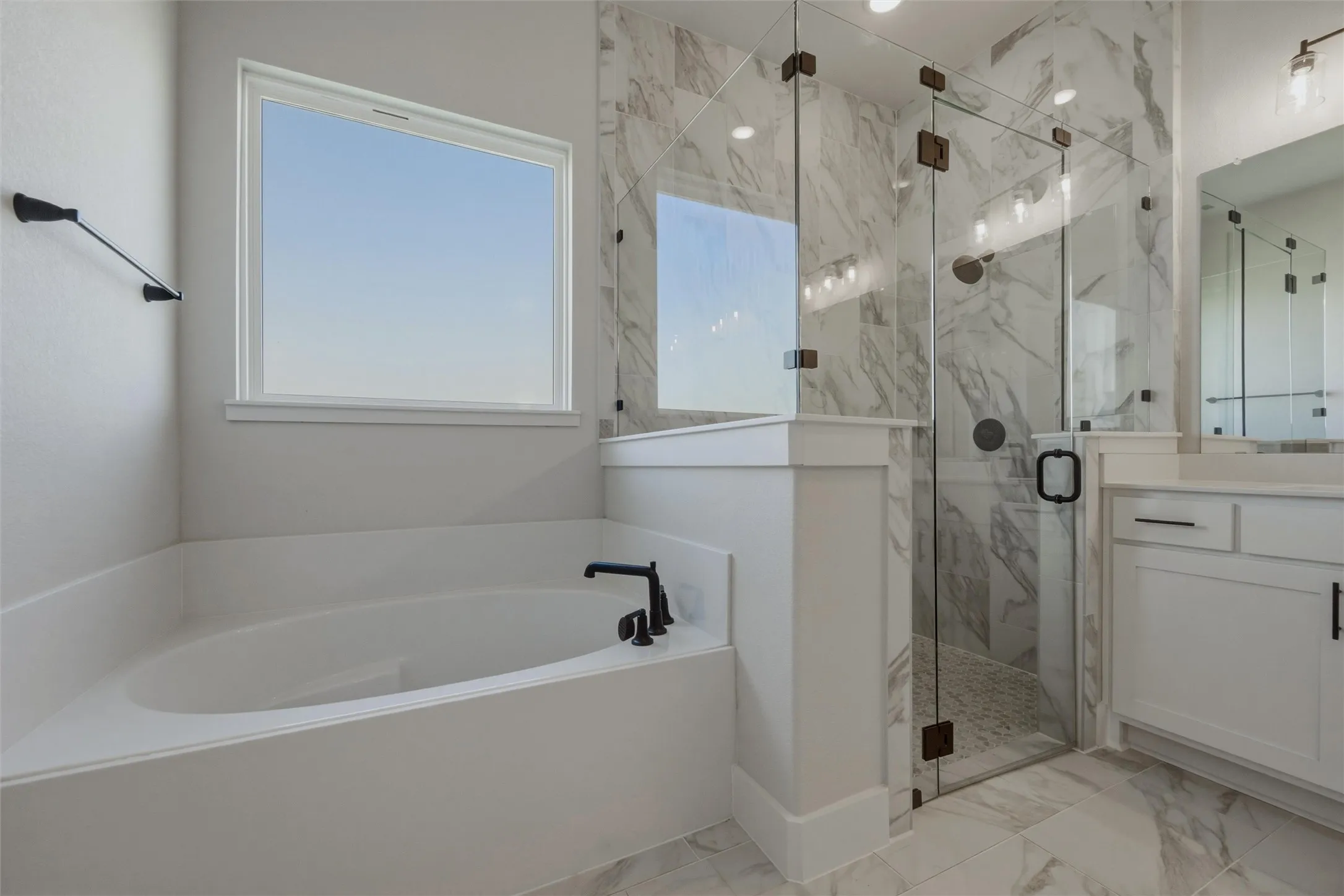 Soaking tub with black fixtures, walk-in shower with clear glass enclosure, white marble-look wall tile, dual vanity with white cabinetry, and large format floor tile