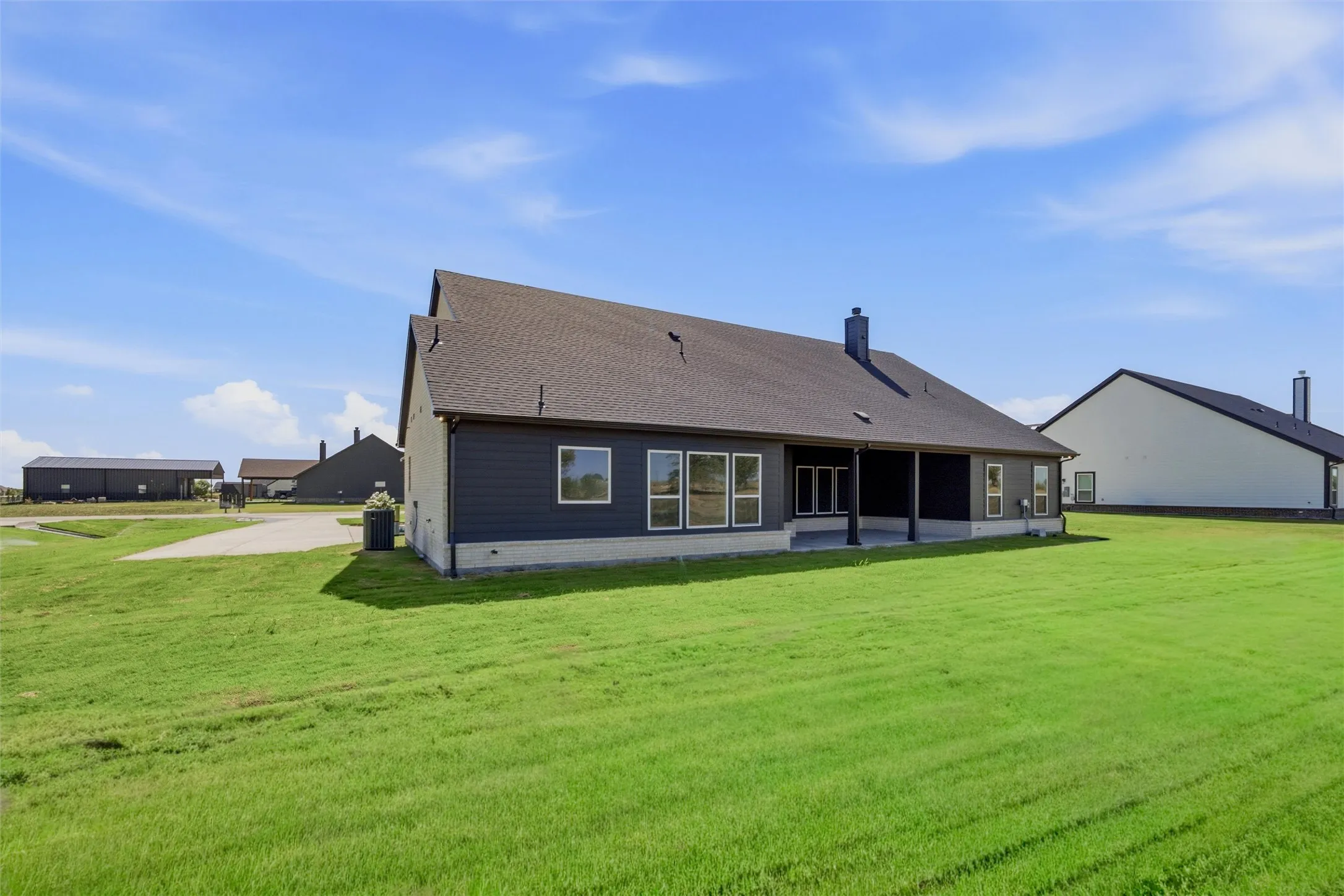 Expansive green space, dark siding with white trim, covered patio, multiple windows, and a dark shingle roof
