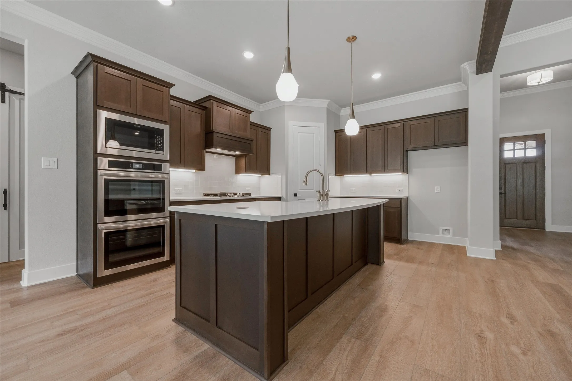 Kitchen featuring a central island with a sink, stainless steel appliances, dark wood-finish cabinetry, light wood-finish flooring, and pendant lighting
