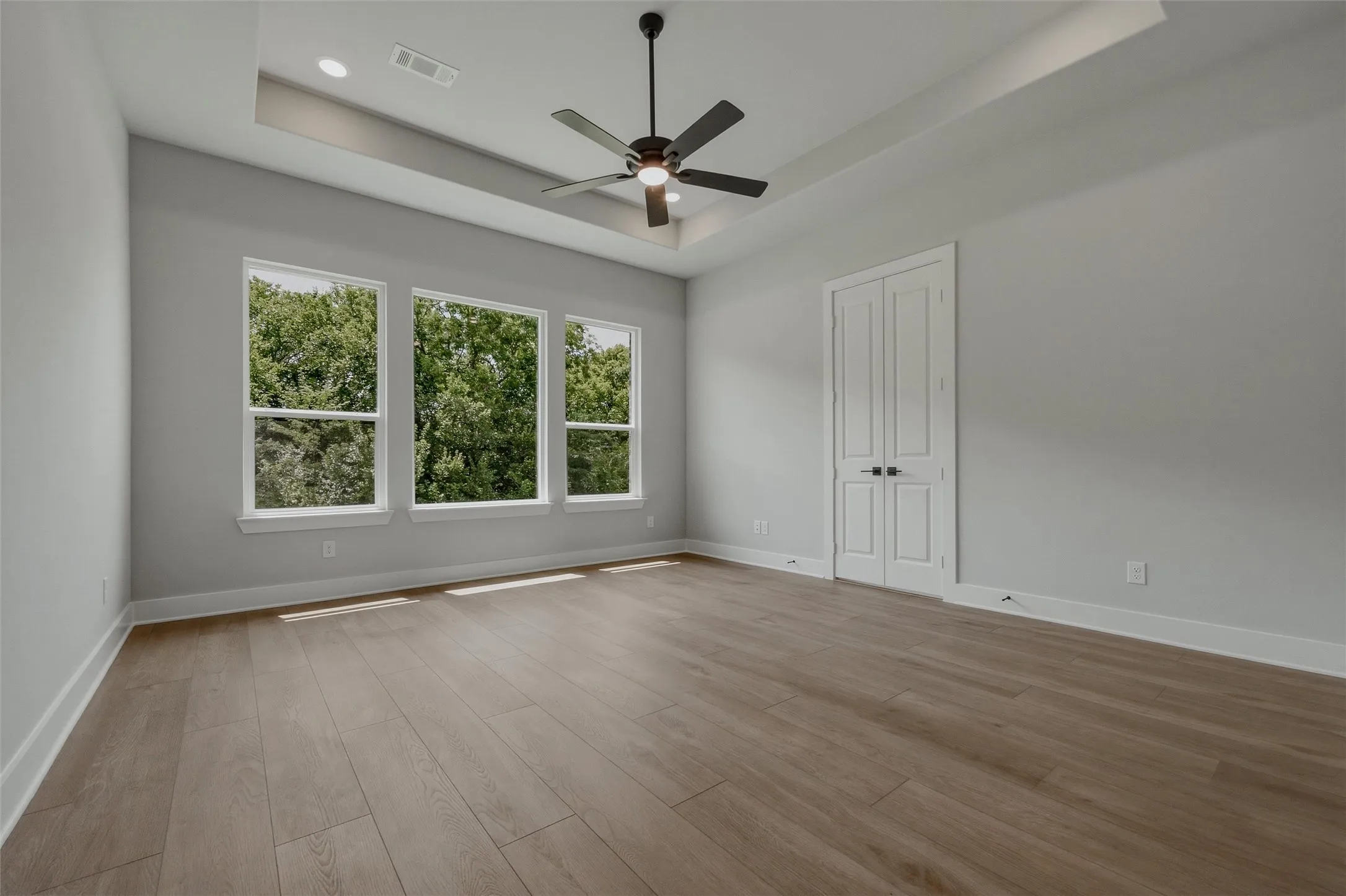 Recessed ceiling with an integrated ceiling fan, three large windows, wood-finish flooring, and white paneled doors with matte black hardware