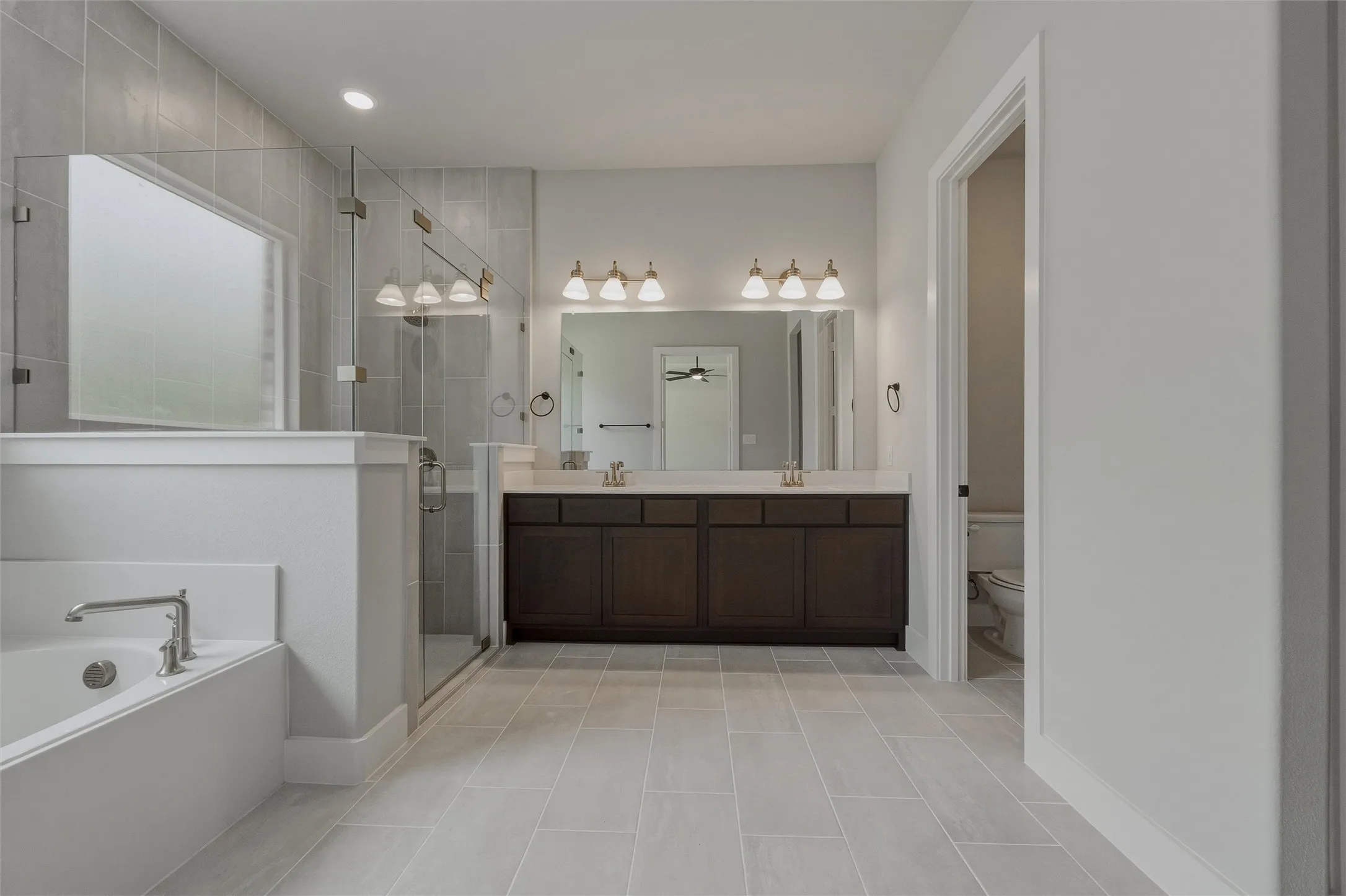 Expansive bathroom featuring light-toned tile flooring, a dual vanity with a dark wood-finish cabinet and light-toned countertop, and a walk-in shower with clear glass enclosure and tiled walls