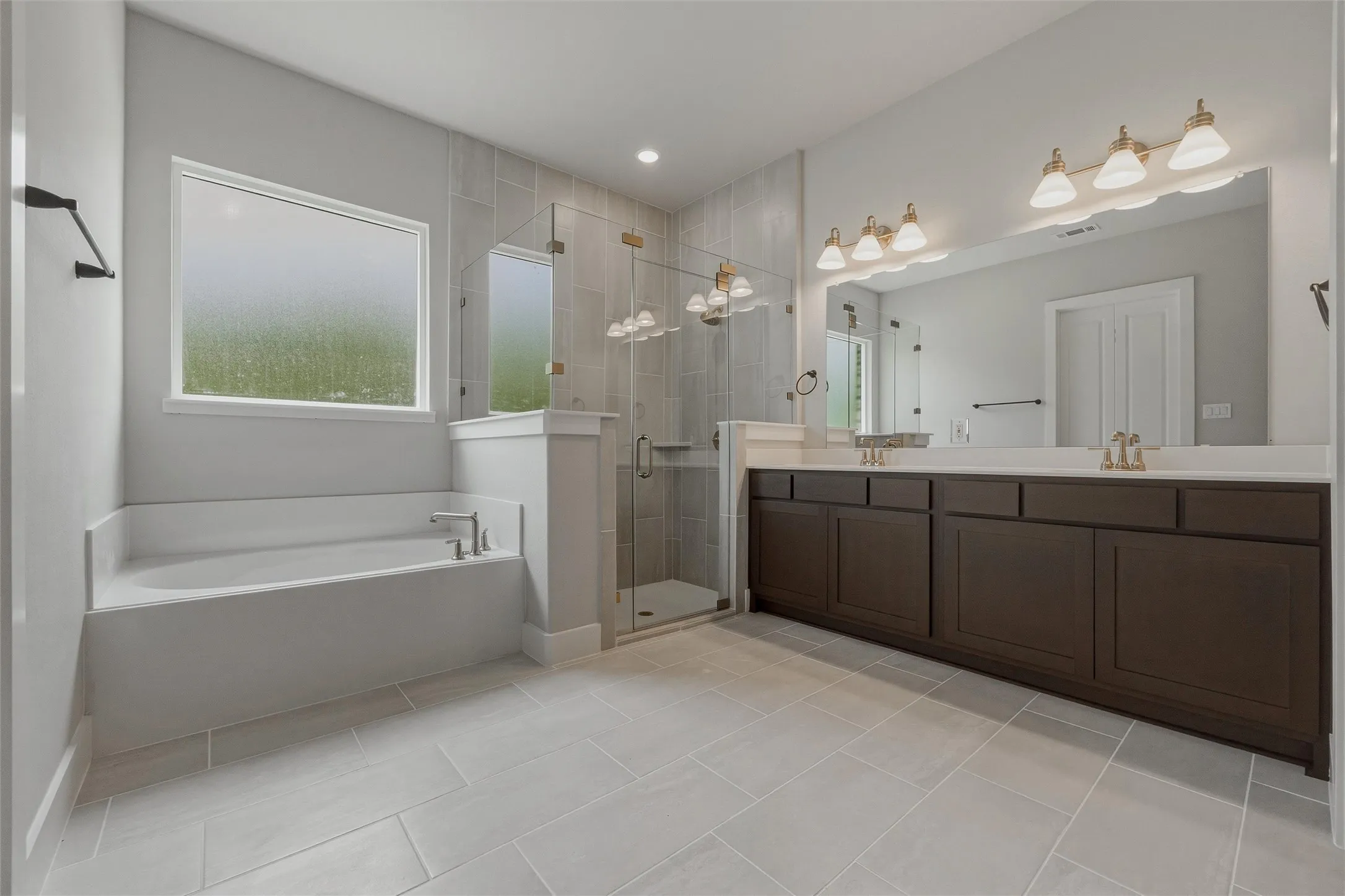 Light-toned tile flooring, built-in soaking tub, walk-in shower with glass enclosure, dual vanity with undermount sinks, and dark cabinetry