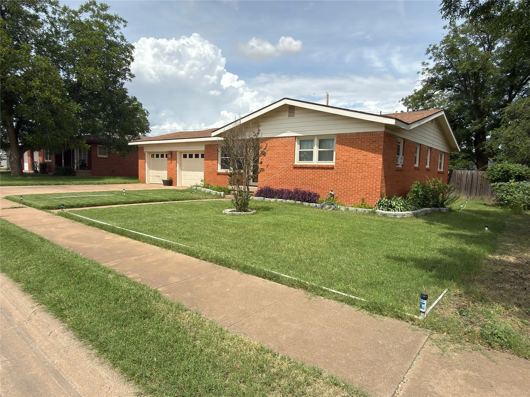 Brick exterior with white trim, featuring a two-car garage with individual doors, established front yard landscaping, and mature trees
