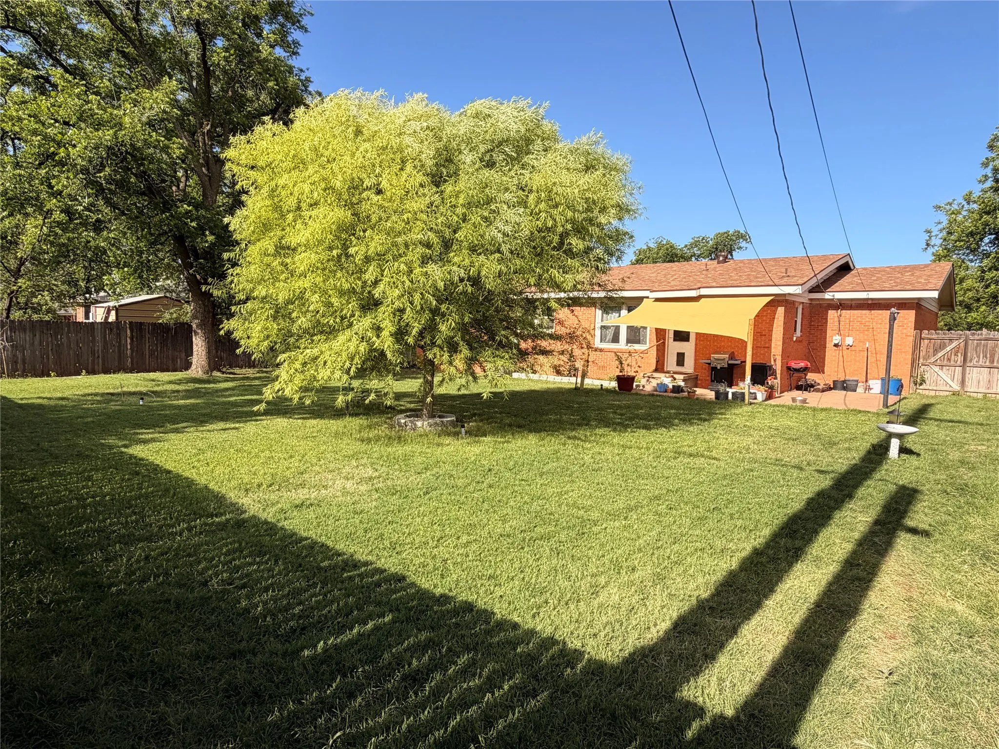 Expansive backyard featuring a large green lawn, mature trees, wood fence perimeter, and a brick exterior dwelling with a covered patio