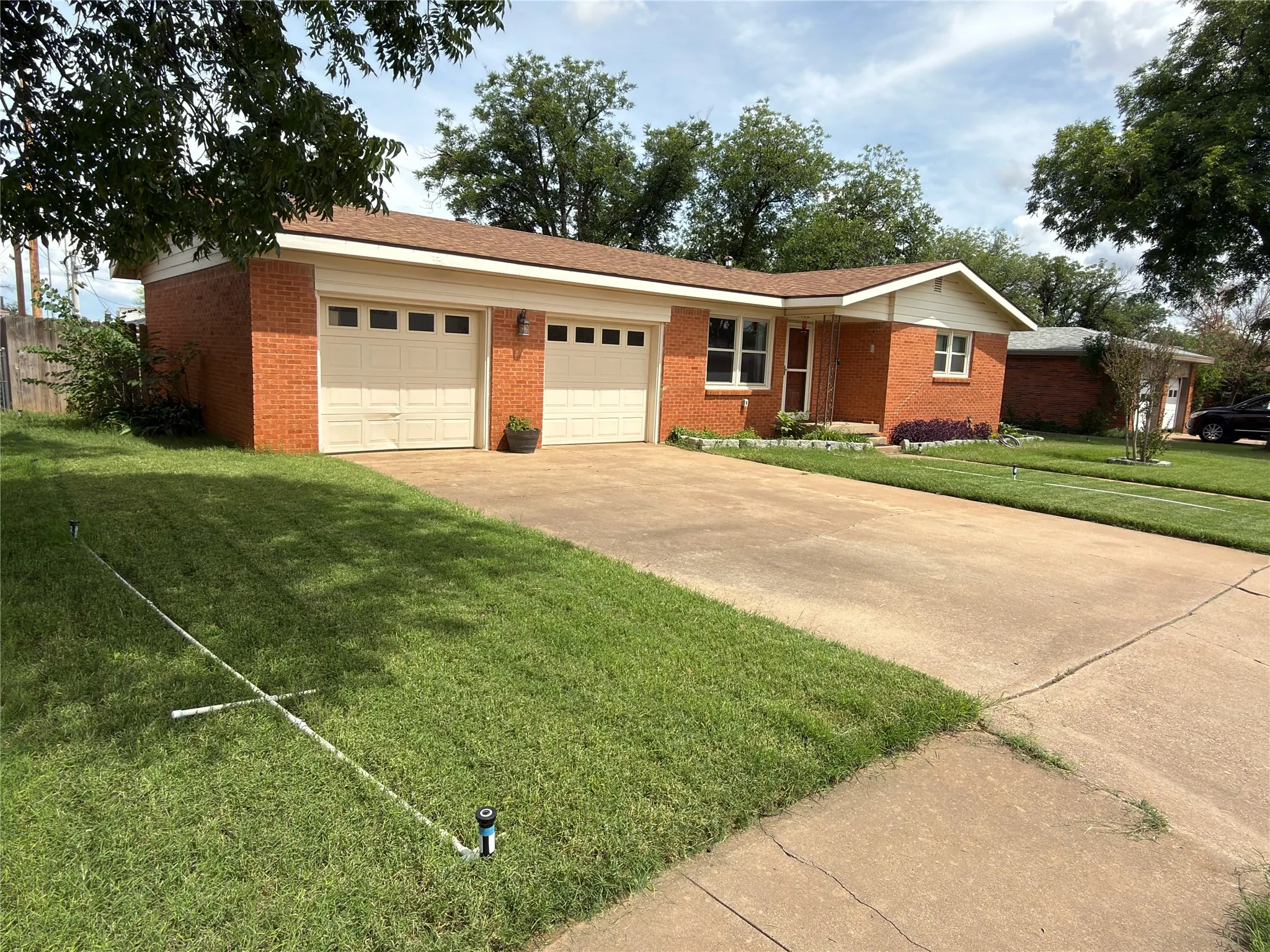 Brick exterior property featuring a two-car garage, concrete driveway, front lawn, and mature trees