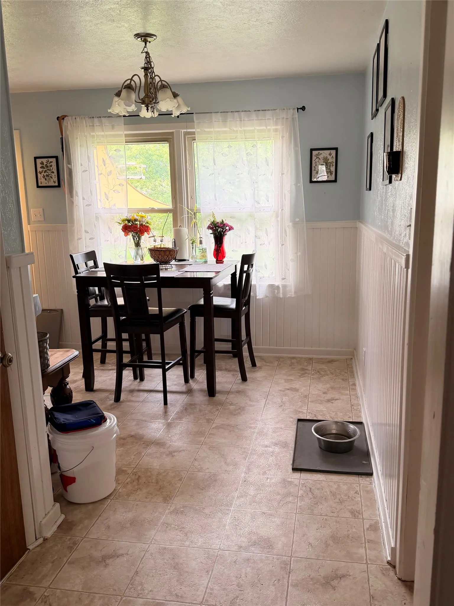 Dining area featuring a central window, overhead chandelier, light blue walls, white beadboard wainscoting, and tile-look flooring