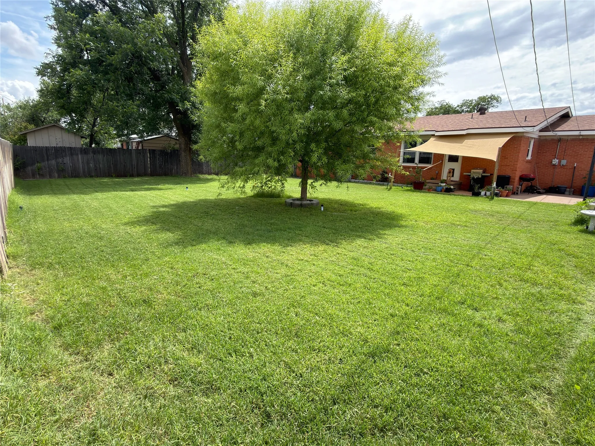 Spacious backyard with a vibrant green lawn, mature shade tree, privacy fencing, and brick exterior with a patio area
