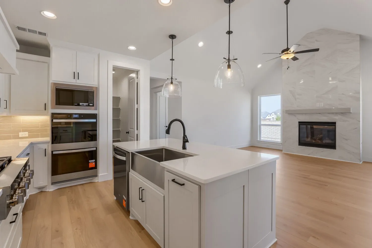 Open-concept kitchen and living area featuring a large central island with a stainless steel farmhouse sink and matte black faucet, light wood-finish flooring, white cabinetry, a built-in stainless steel microwave and double oven, and a tall fireplace with a natural stone surround and mantle