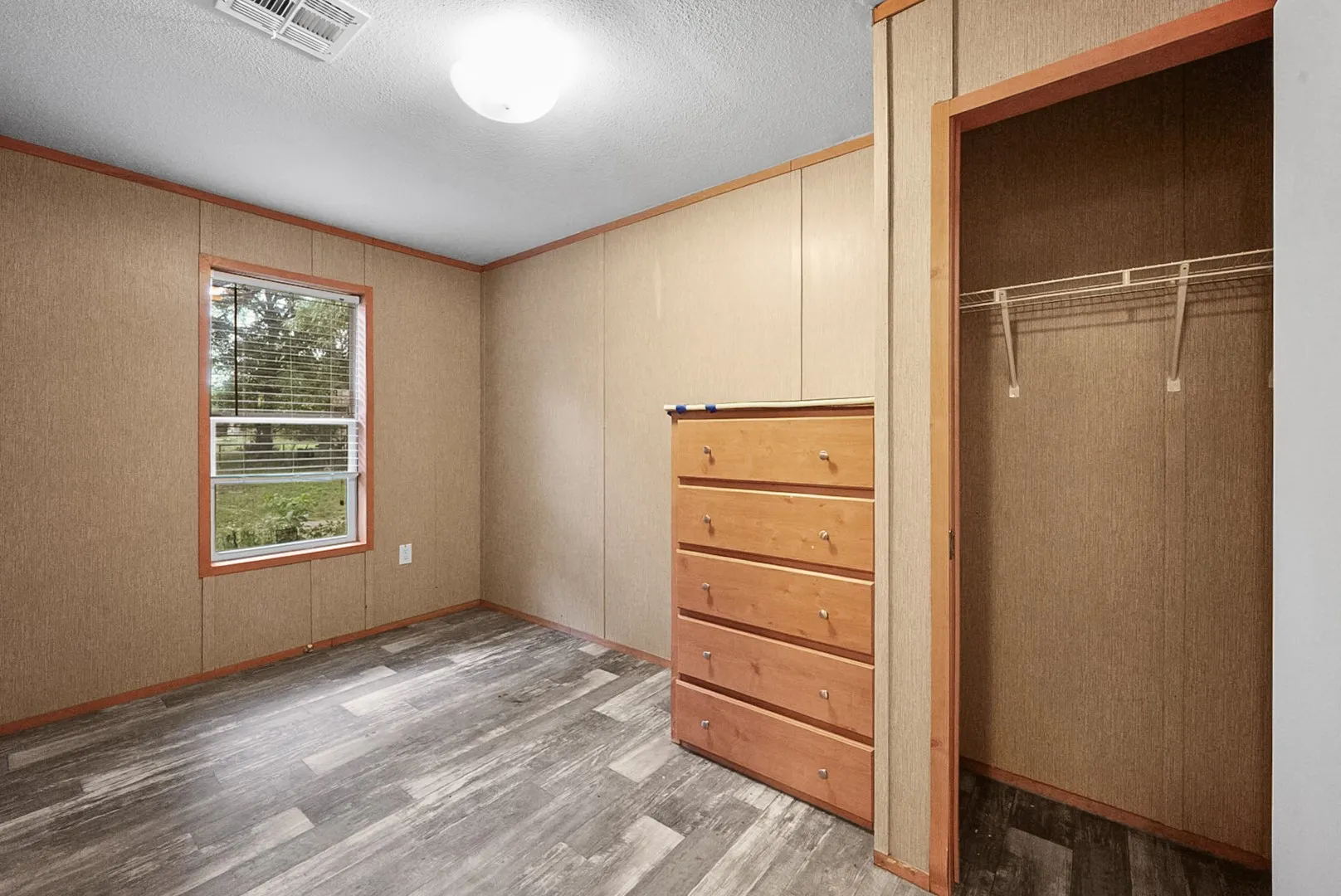Room featuring wood-finish flooring, light-toned wall paneling, and a single window with blinds