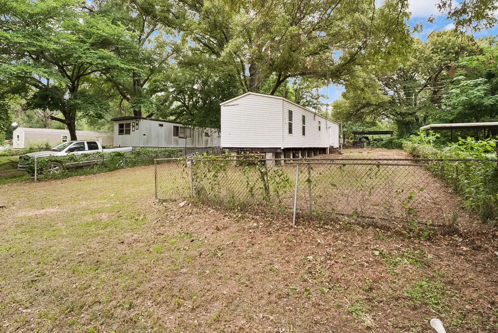 Chain-link fenced property featuring two white mobile homes, mature trees, and an additional covered structure