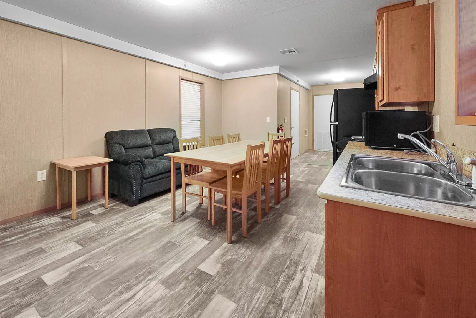 Open-concept living space featuring wood-finish flooring, a kitchen with wood-finish cabinetry, a double basin stainless steel sink, and light-toned countertops